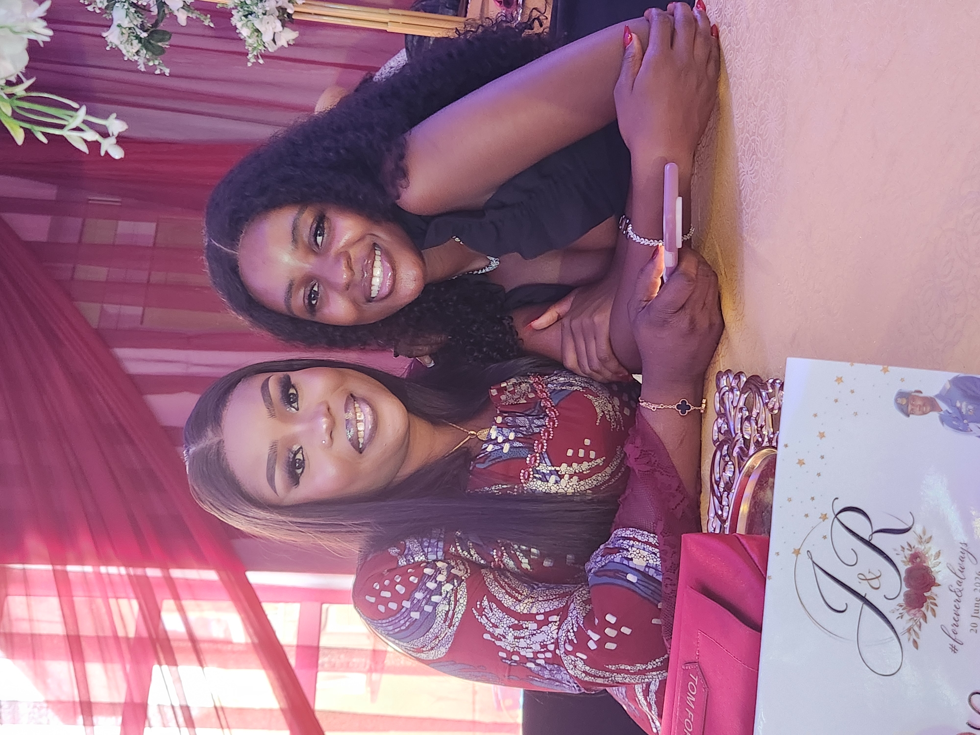 Two women are seated at a table, posing for a photograph. The woman on the left wears a red and gold patterned dress and has her hair pulled back. She is smiling, and her makeup is visible. The woman on the right has long, dark, curly hair and wears a black top. She is also smiling broadly. Both women have jewelry on their wrists. In the foreground, a red clutch bag and a white card with "J&R #forever&always" printed on it are visible. The background features red drapes and white floral arrangements. The overall setting appears to be an indoor event space, possibly a reception hall, in Barden Yana, Nigeria, suggested by the cultural attire and the event invitation.