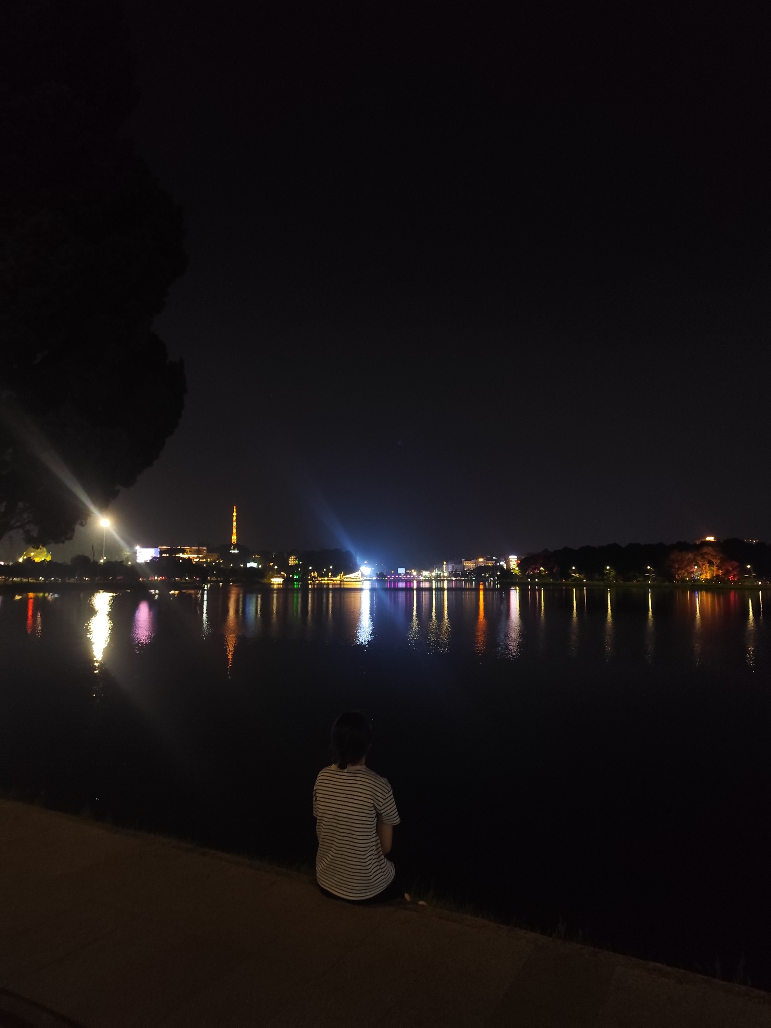A person sits by the edge of Ho Xuan Huong Lake in Da Lat, Vietnam, at night. The person has short dark hair and wears a striped white and dark blue shirt. They face the lake and the city lights that reflect in the water. In the background, a tower is lit orange and white. Further down the shoreline are other lights, white, blue, and orange, of various shapes and intensities. A large dark tree is on the left side of the image. The pavement where the person sits is lit by ground-level light. The sky is dark.