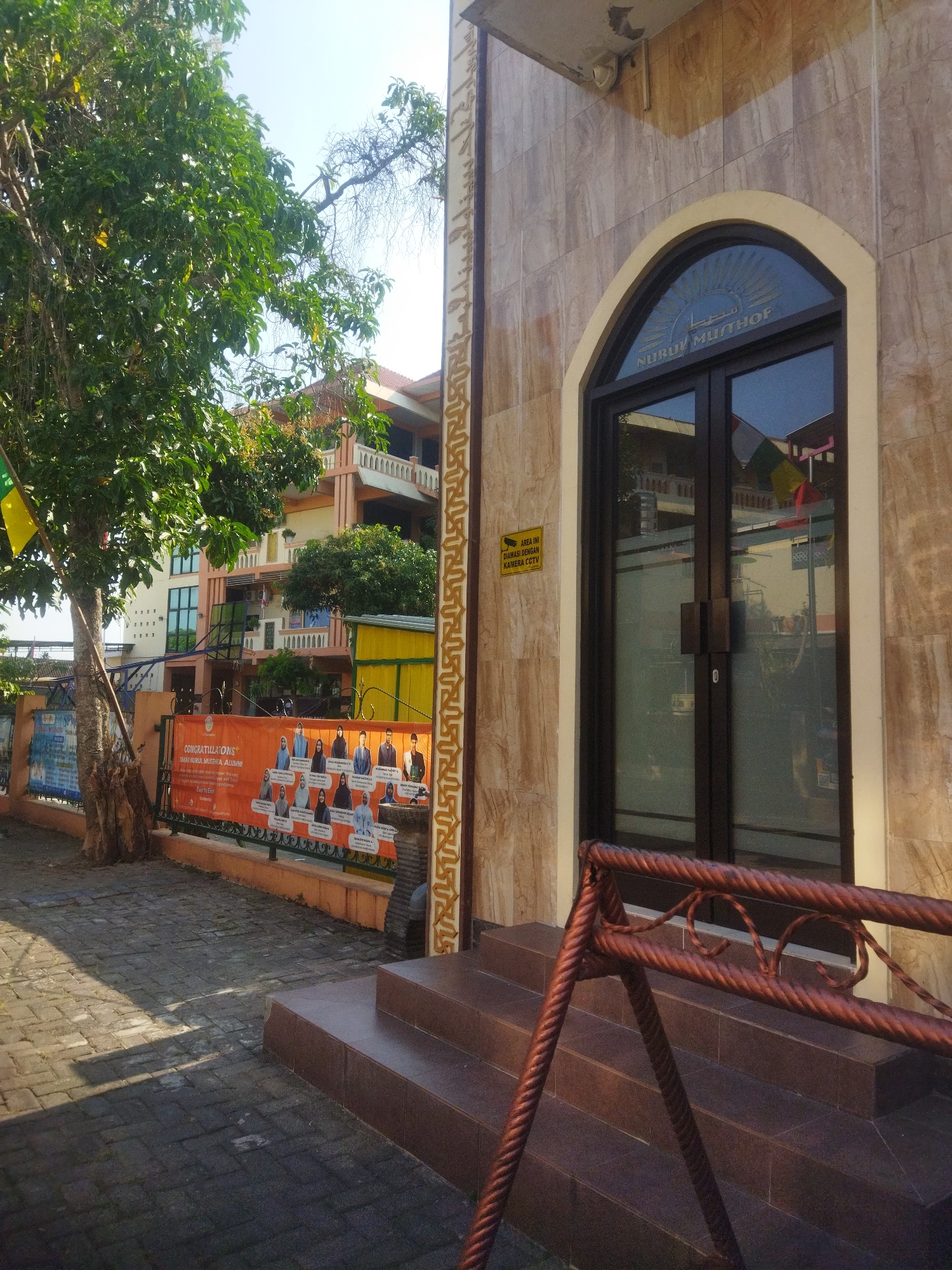 The image shows the exterior of a building in Juwiring, Indonesia, with a large tree on the left side of the frame. The building is made of light brown marbled tiles and features a large arched doorway with double glass doors. Above the doors, Arabic script is visible, along with a sunburst design. A yellow sign indicating CCTV surveillance is posted to the left of the doorway. In front of the building, there are several steps leading up to the entrance. To the left of the building, there is a sidewalk paved with dark grey bricks. Further to the left, a fence is visible with an orange banner displaying a group of people. Behind the fence, there is a multi-story building with balconies and windows. The sky is clear and blue. The overall scene depicts a sunny day in an urban or semi-urban environment.