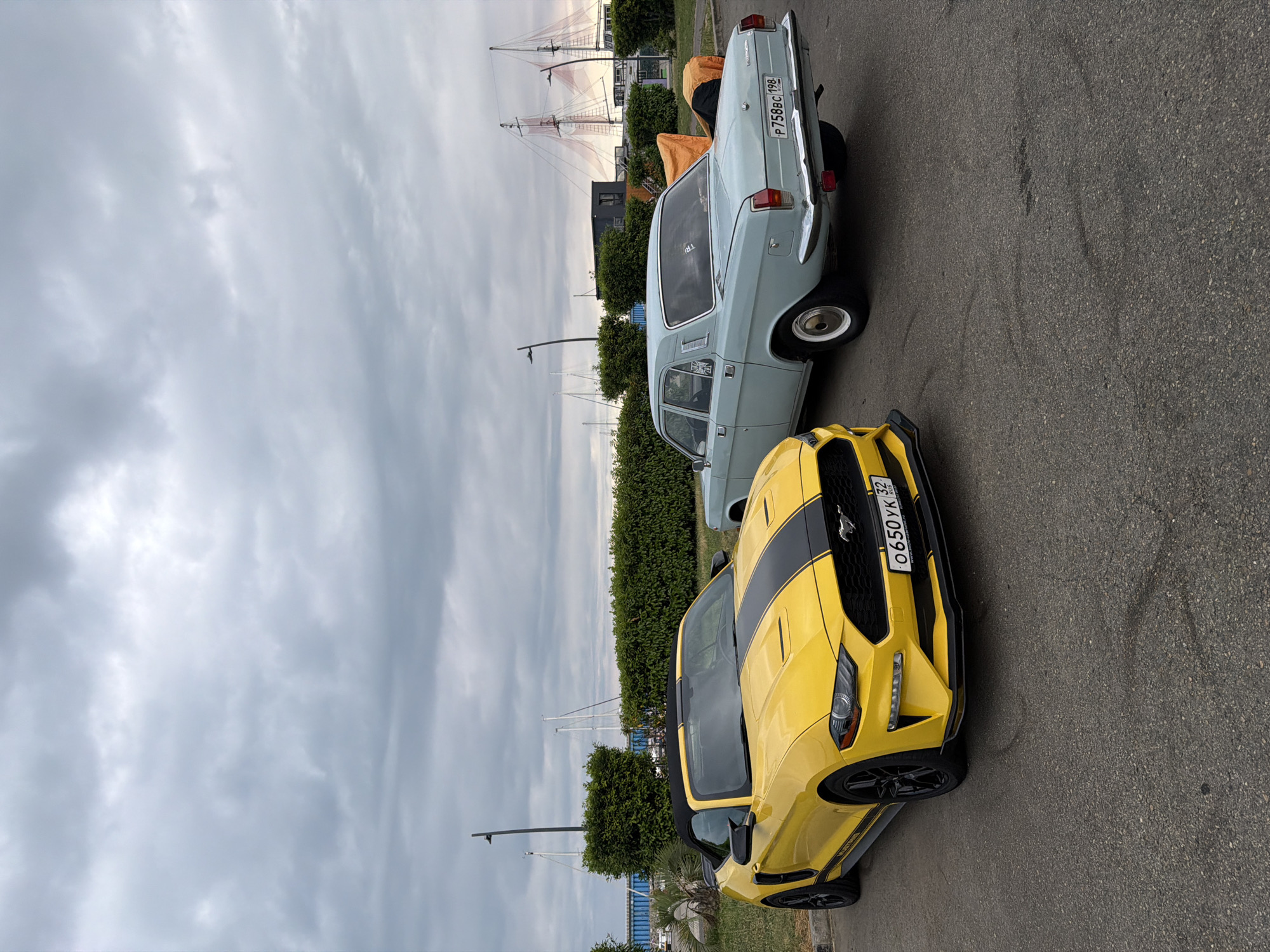 Two cars are parked on asphalt, with a cloudy sky overhead. The car in the foreground is a yellow convertible Ford Mustang with black racing stripes. Its license plate reads "O 650 YK 32 RUS". The car in the background is a light blue, older model sedan, possibly a GAZ-24 Volga, with a license plate reading "P 758BC 198". In the background, there are manicured green bushes, a body of water with sailboats, and masts of ships, suggesting a waterfront location. The overall scene appears to be a parking area near a harbor or marina in the city of Sirius, Russia.