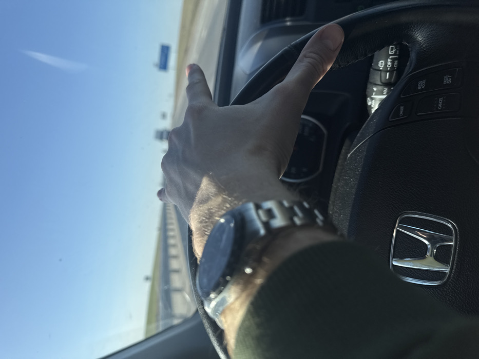 The image displays a first-person perspective from the driver's seat of a Honda vehicle in Абухаўскі сельскі Савет, Belarus, while in motion. The driver's right hand, wearing a metallic smartwatch on the left wrist, grips the upper right portion of the black leather steering wheel, which features a silver Honda emblem in the center and cruise control buttons on the right spoke. The driver is clad in a dark green long-sleeved garment. Ahead, a multi-lane highway stretches into the distance under a clear blue sky, containing a faint aircraft contrail. A metallic guardrail lines the left side of the road, and distant vehicles along with a blue rectangular road sign are visible further down the highway. The scene suggests a sunny day with unimpeded forward visibility.