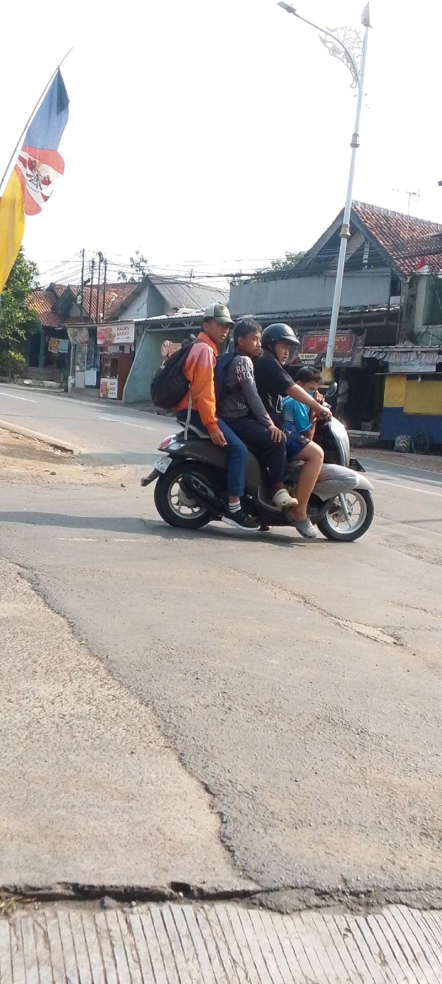 The image shows a street scene in Cilaku, Indonesia, with four young individuals riding on a single motorcycle. The motorcycle is a dark, possibly grey, scooter. The rider in the front is wearing a blue shirt and dark shorts, with a helmet. Seated behind him is a person in a black t-shirt and dark pants. Next is a person in a grey long-sleeved shirt and dark pants, with a backpack on their back. The person furthest back is wearing an orange jacket and dark pants, also with a backpack. They are all looking forward, suggesting they are traveling on the road. The road appears to be asphalt and shows signs of wear, with some cracks. On the right side of the road, there are buildings with tiled roofs, and some commercial stalls are visible. A street light with a decorative design is also present. On the left side of the image, a flag with red, blue, and yellow colors is flying. The sky is bright, indicating daylight.