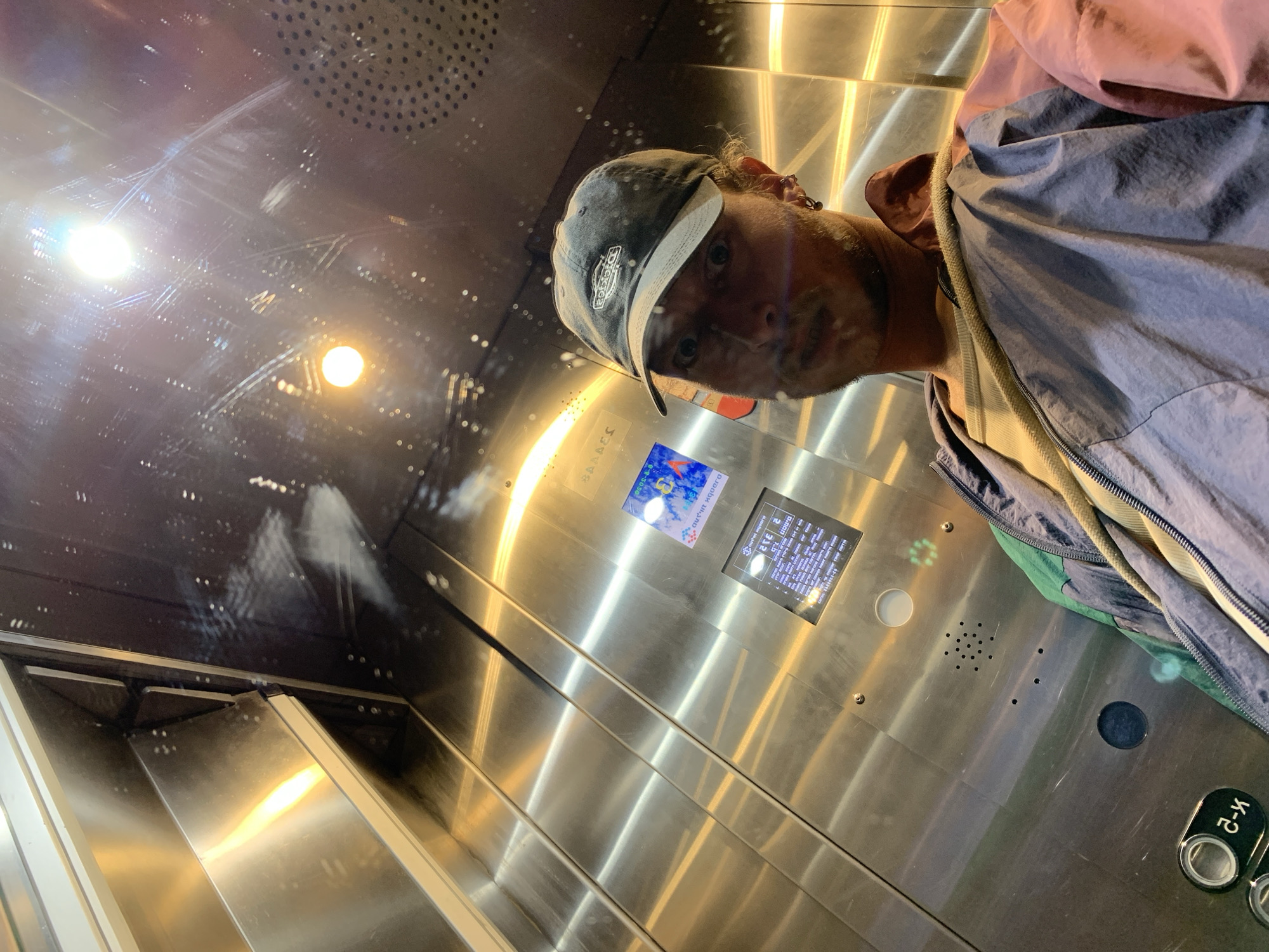 The image shows a man taking a selfie in an elevator. The elevator has a stainless steel interior. The man is wearing a baseball cap and a blue and pink jacket. He appears to be looking into the camera.
The elevator's control panel is visible, along with an advertisement card. The button panel includes Braille labels.
The reflection in the elevator shows the interior ceiling and lights. The elevator is located in Tel Aviv, Israel, based on the Hebrew writing on the advertisement card.
