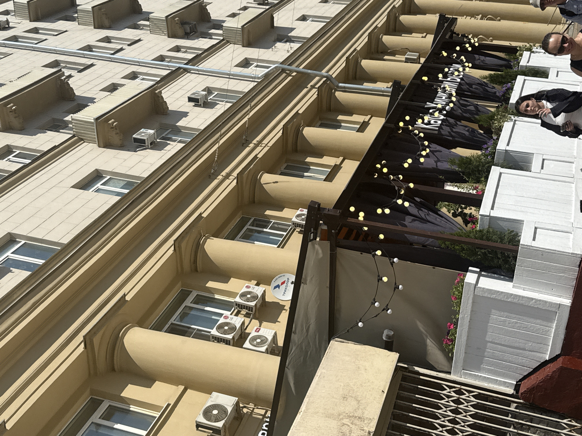 This is an eye-level shot of a tan building in Moscow, Russia. The building is tall and several stories high, with rows of windows and air conditioning units attached to the exterior walls. The building has a classical architectural style with columns and decorative molding.
In the foreground of the image, there is an outdoor patio area. The patio has a metal awning structure with hanging string lights. Below the awning are dark-colored sun umbrellas, tables, and white planters with plants and flowers. People are standing and walking around the patio area.
The overall environment appears to be a sunny day, and the building is located in an urban setting.