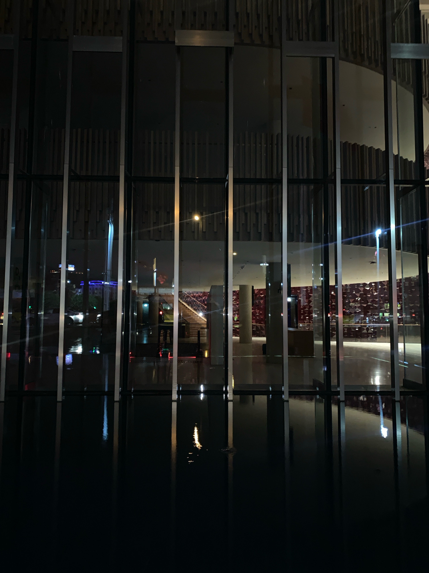 This is a nighttime exterior shot of a modern building with a glass facade, possibly in Beyoğlu, Turkey. The building features vertical metal framing and wooden slats on the upper portion of the facade. The glass reflects the street scene at night, including lights from vehicles and buildings, and what appears to be a body of water in the foreground, reflecting the building and the external lights. Inside the building, visible through the glass, are architectural elements such as columns, a staircase with red accents, and walls covered in a faceted red pattern. There are no people clearly visible in the image. The overall atmosphere is dark and reflective, with the city lights creating streaks and reflections on the wet-looking surface in front of the building.