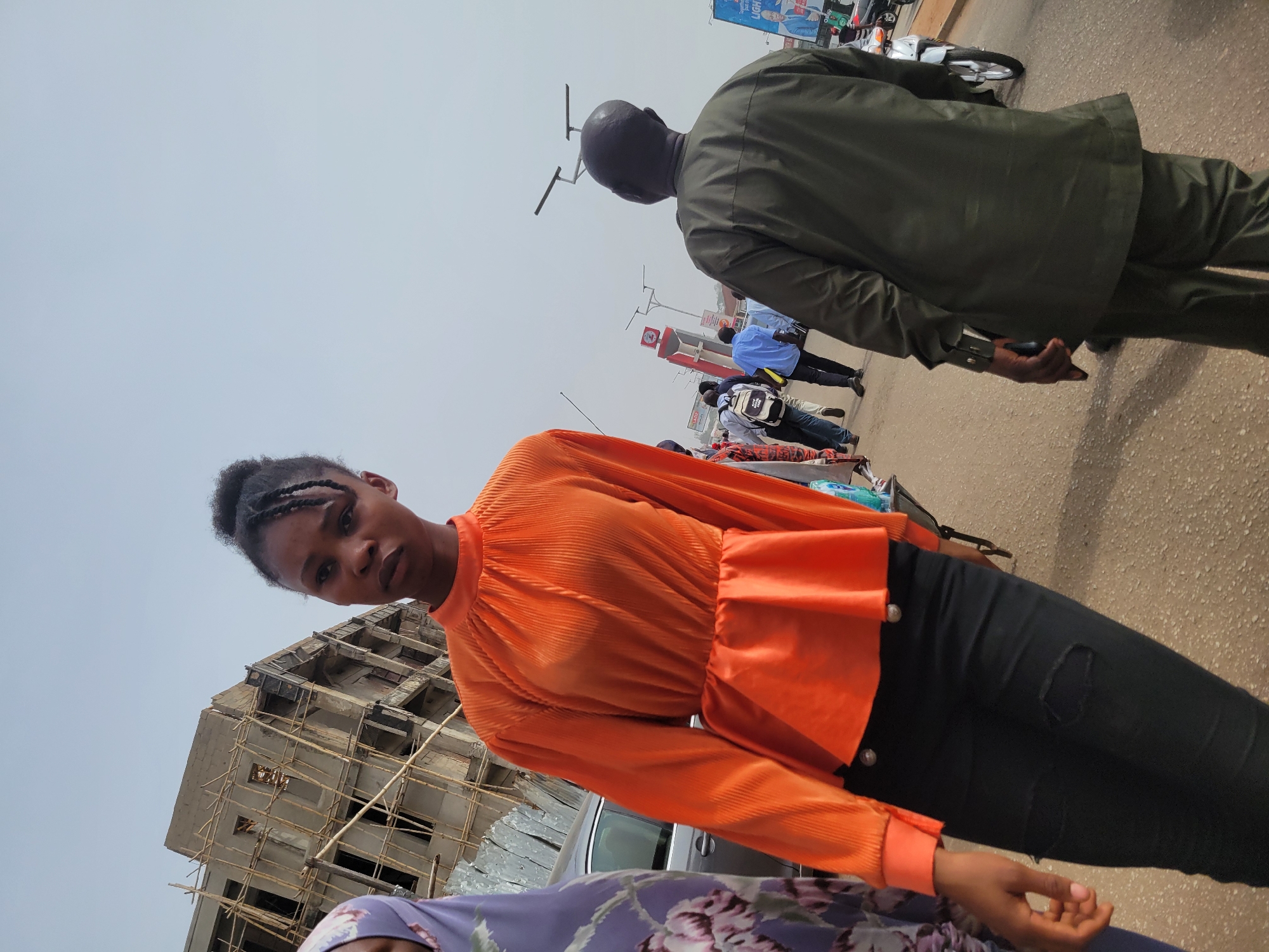 The image captures a bustling street scene in Jos, Nigeria, under what appears to be a bright, hazy sky. The primary subject is a young woman, positioned prominently in the foreground, looking directly at the camera with a neutral expression. She is wearing a vibrant orange, long-sleeved top with a pleated texture and ruffled detailing around the waist, paired with black jeans that have a rip on the left thigh. Her hair is styled in dark braids.
Partially visible in the lower left corner is another person wearing a purple garment with a floral pattern. In the mid-ground, a man in a dark green two-piece suit is walking away from the camera, his back to the viewer. Several other pedestrians are scattered further down the street, and a silver car is partially visible behind the main subject. A motorcycle is also seen in the background.
To the left of the main subject, a multi-story building is under construction, featuring a concrete frame surrounded by extensive wooden scaffolding. The street appears to be asphalt, and various commercial buildings or structures line the background. In the far distance, a billboard displays the partially visible word "LIGHT." The scene suggests an active urban or semi-urban environment with ongoing development during the daytime.