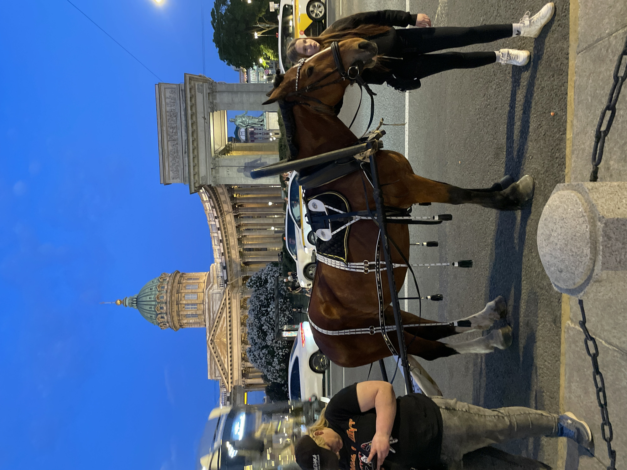 The image captures an urban twilight scene in Saint Petersburg, Russia, centered on a horse and two individuals, with prominent architectural landmarks in the background.
**Location:** The scene is set on a street fronting the Kazan Cathedral and the General Staff Building. The Kazan Cathedral, identifiable by its large green dome with a gold finial and a classical colonnade, occupies the left-center background. To its right, the Triumphal Arch of the General Staff Building is visible, featuring sculpted relief panels and revealing a statue of a standing male figure through its opening.
**Foreground Subjects:**
* A brown horse, equipped with a black and white harness and bridle, stands facing slightly right. The harness features several decorative white and black tassels hanging down its side. The horse has four white markings on its lower legs (socks/stockings) and a dark mane.
* A young female stands to the right of the horse, holding its bridle with her right hand. She has long, dark hair and wears a black hooded sweatshirt, black leggings, and white athletic shoes with white socks. She is looking towards the horse's head.
* An adult female stands to the left, partially obscured by a reflective surface, possibly a vehicle. She wears a black t-shirt with a graphic design featuring stylized faces and Cyrillic text, grey jeans, and blue and white athletic shoes. She also wears a black baseball cap with partially visible Cyrillic text. She is holding a smartphone with both hands and appears focused on its screen.
**Midground Elements:**
* An asphalt street separates the foreground subjects from the background buildings.
* Several vehicles are present on the street: a white luxury sedan (e.g., Porsche Panamera-like) is prominent in the mid-left background, and a white and yellow taxi is visible further to the right.
* A large tree with white blossoms stands behind the white sedan, partially obscuring the lower part of the General Staff Building.
* In the bottom-right foreground, a gray stone pillar with a black metal chain barrier is visible.
**Environmental Details:** The sky is a clear, deep blue, indicative of twilight or early evening. Artificial lighting illuminates the architectural landmarks and distant streetlights are visible. The ground consists of asphalt pavement with yellow painted lines and a lighter-colored sidewalk.