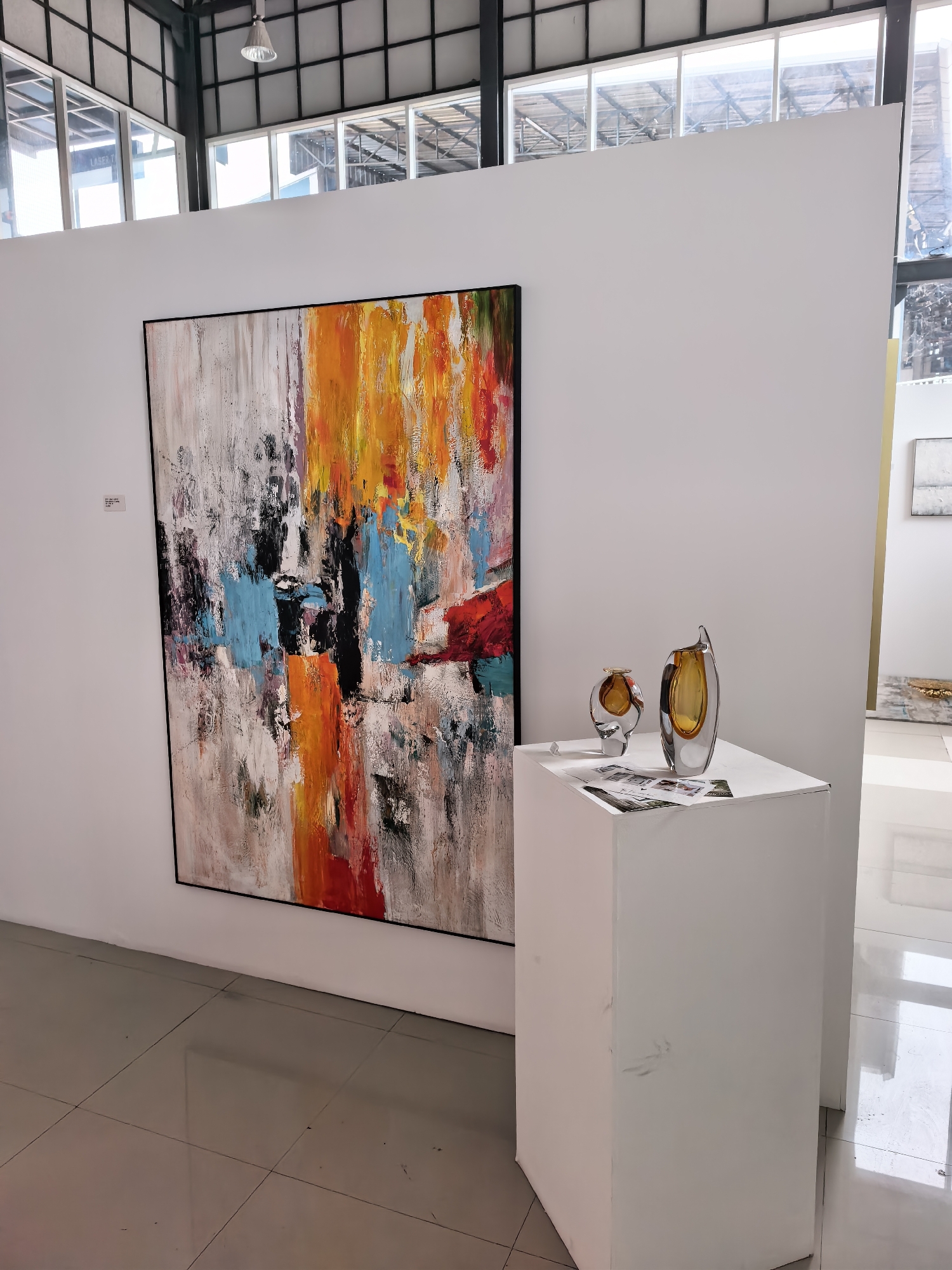 The image shows an interior art gallery or exhibition space, possibly in the city of Ko Samui, Thailand, given the architectural style and lighting. The primary focus is a large abstract painting on a white wall, featuring bold strokes of orange, yellow, red, blue, and black against a textured white and gray background. To the right of the painting, a white pedestal displays two modern glass vases with amber-colored interiors. The floor is tiled with large, light-colored square tiles that reflect the overhead lighting and the surrounding environment. Large windows with metal framing are visible in the background, suggesting a spacious, well-lit venue. The overall impression is one of contemporary art display.