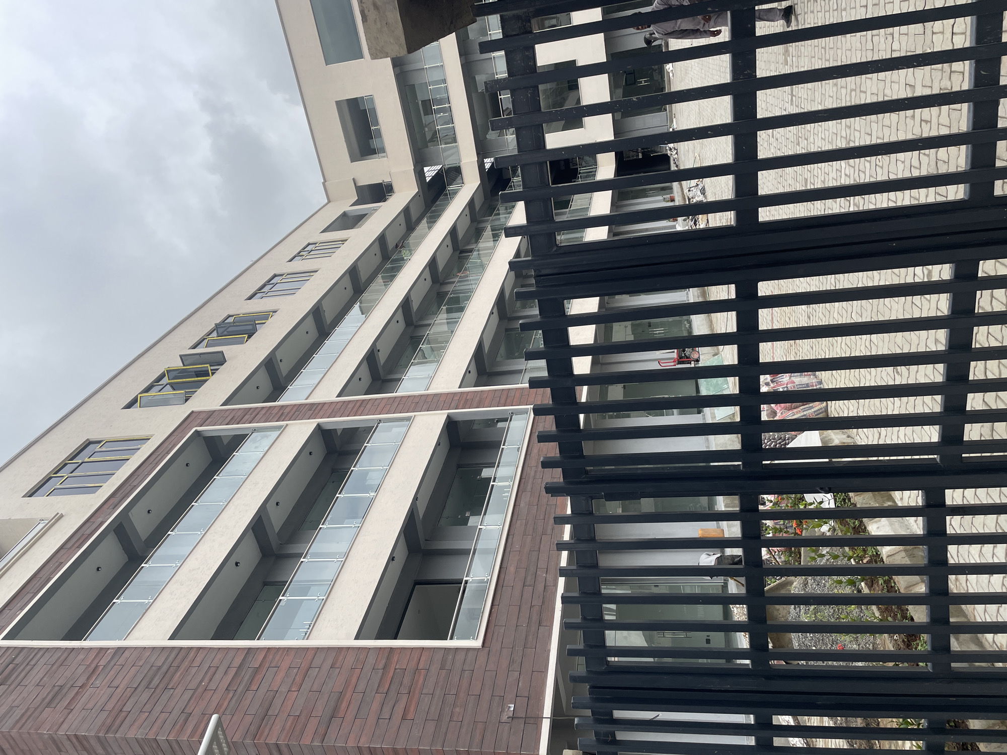 This is a photograph of a modern, multi-story building under construction in Abuja, Nigeria. The building features a light-colored facade with dark brick accents on the left side. Several floors have balconies with glass railings, and there are numerous windows, some with yellow frames. The building appears to be part of a larger complex, with an open courtyard visible through a black metal fence in the foreground. The courtyard shows ongoing construction work, with piles of gravel, some bagged materials, and a few small plants. Two individuals are visible in the courtyard, one appearing to be working and the other standing near the fence. The sky is overcast, suggesting a cloudy day.