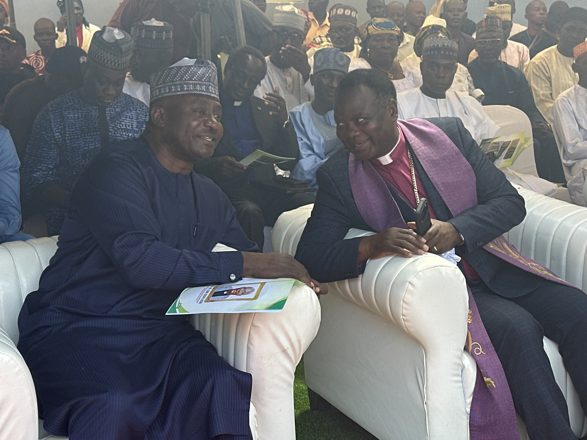 The image depicts a formal gathering in Sabon Tasha, Nigeria, featuring two prominent male figures seated on white upholstered armchairs in the foreground, engaging in conversation. Behind them, a large group of people, predominantly men, are seated in rows, forming an audience.
The man on the left is of dark complexion, wearing a dark blue garment with subtle vertical stripes, consistent with traditional Nigerian attire. He has a light blue cap with a geometric white and black pattern on his head. He is smiling broadly and looking towards the man on his right. His right hand rests on a green and white pamphlet or program, which displays a framed portrait of a man in a clerical collar.
The man on the right, also of dark complexion, is wearing a dark suit over a white clerical shirt with a white collar. A purple stole, adorned with gold embroidery, is draped around his neck and over his shoulders. He is smiling and looking at the man on his left, with his head slightly tilted. His hands are clasped or resting on a dark, handheld object, possibly a mobile phone or small microphone, in his lap.
Both men appear to be in their middle to older adult years. Their interaction suggests a friendly and engaged conversation.
The background shows numerous attendees, most of whom are men, seated and observing the scene. Many are dressed in various forms of traditional Nigerian clothing, including different styles of caps and robes, while some wear Western attire. A few women, also wearing head coverings, are visible among the crowd. The clarity of individuals in the background diminishes with distance.
The foreground subjects are seated on modern, plush white armchairs, which are placed on what appears to be green artificial turf or a short-pile green carpet. The lighting is bright and even, illuminating the scene clearly. The overall setting indicates an organized event or assembly.