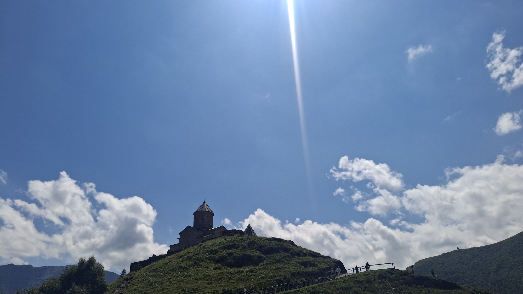 The image shows Gergeti Trinity Church, a 14th-century Orthodox church located on a steep mountain slope in Stepantsminda, Georgia. The church, made of stone, has a distinct conical roof and a cross on top. It is situated on a green, grassy hill against a backdrop of a bright blue sky with scattered white clouds. The sun is visible, creating a strong lens flare that streaks down from the top right of the frame. Several people are seen walking up a path towards the church, some silhouetted against the sky. In the distance, other mountain peaks are visible. The environment appears to be mountainous and natural.