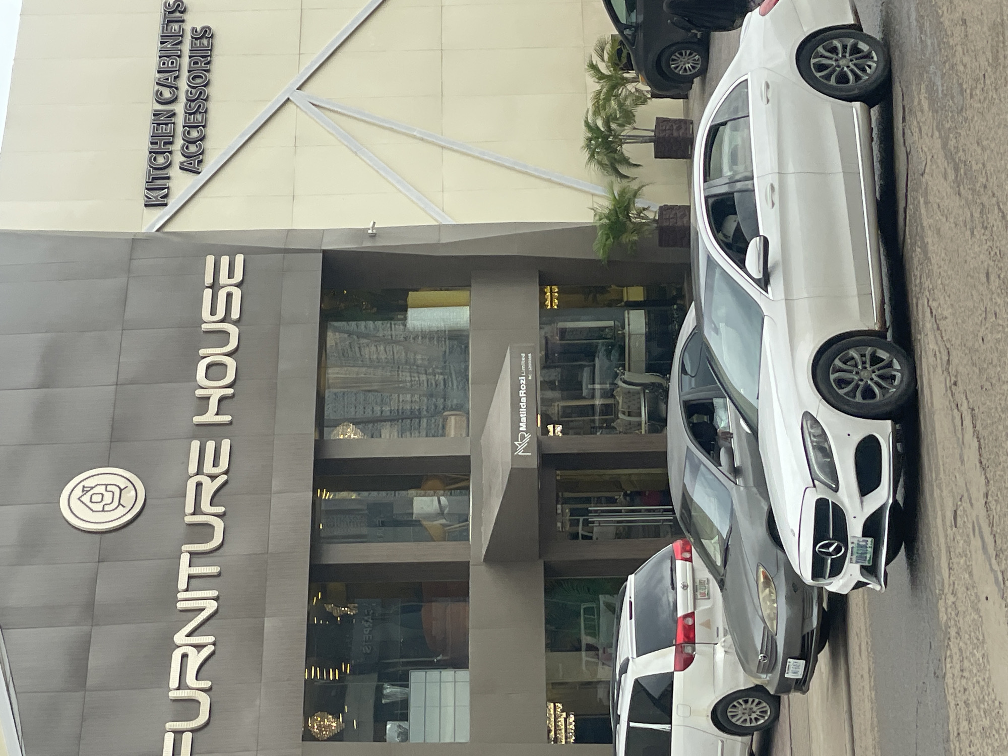 The image shows the exterior of the "FURNITURE HOUSE" store and "KITCHEN CABINETS ACCESSORIES" in Abuja, Nigeria. In the foreground, three cars are visible on a paved road. The closest car is a white Mercedes-Benz sedan, followed by a grey sedan and a white minivan. The buildings have a modern architectural style with large glass windows showcasing interior furniture. The environment appears to be a commercial area with potted palm trees outside the buildings.