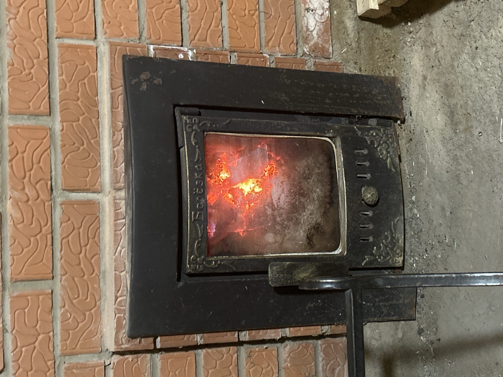 The image shows a close-up of a black, ornate, rectangular metal wood-burning stove set into a red brick wall. Flames are visible through the dirty glass door of the stove. The stove has a handle and several small vents on the right side of the door. The environment suggests an indoor setting. The location in the city of Уральское, Russia, cannot be confirmed based on visual information alone.