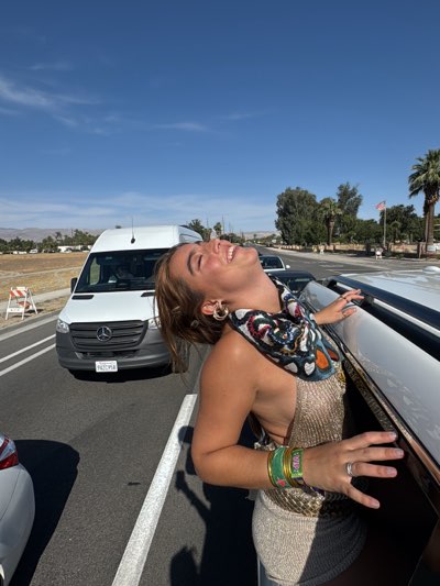 A woman is leaning out of the open roof of a white vehicle with a black stripe on a bright sunny day. She has her head tilted back and is smiling. She is wearing a shiny gold halter top dress, hoop earrings, colorful bracelets, and a patterned scarf. A white van is stopped in front of the vehicle with the woman, and there is a white car to her left. Palm trees and green vegetation are visible in the background, along with distant mountains. A United States flag is visible on a flagpole in the background. The license plate of the van is visible, with the number 9427958. The image was taken in the city of Indio, United States.