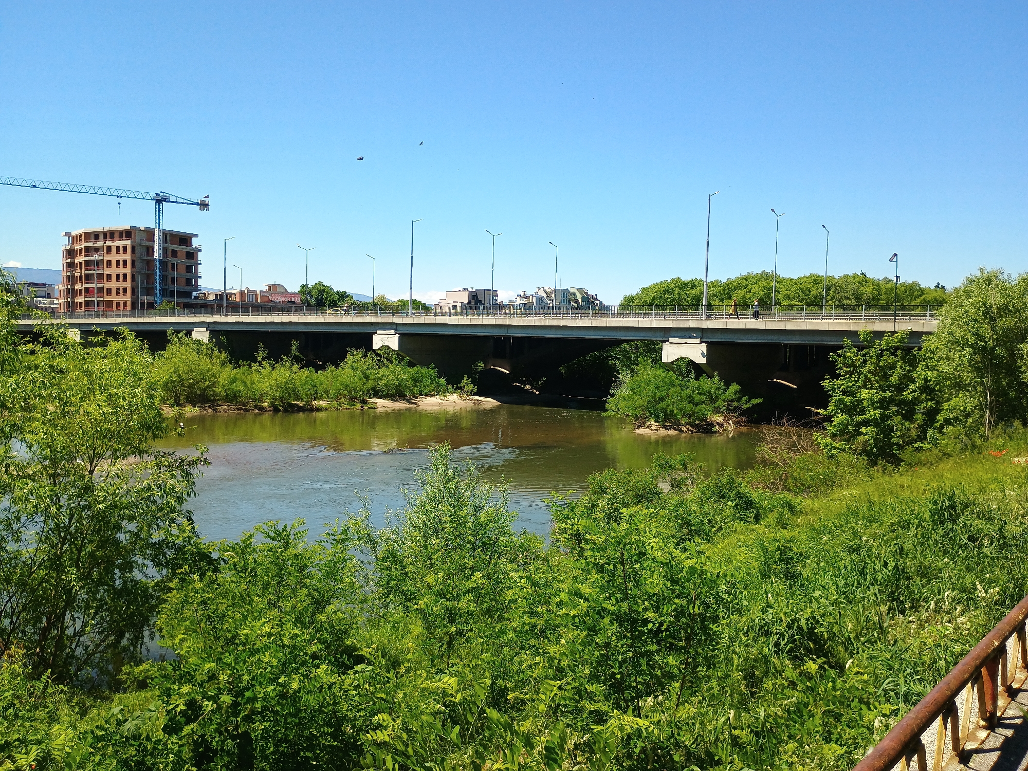The image depicts a bridge spanning a river, with lush green vegetation in the foreground and background. In the distance, a building under construction is visible, with a blue crane positioned next to it. The bridge is populated with street lights and appears to carry vehicle and pedestrian traffic. The sky is clear and blue. The location is in the city of Unknown, Unknown.