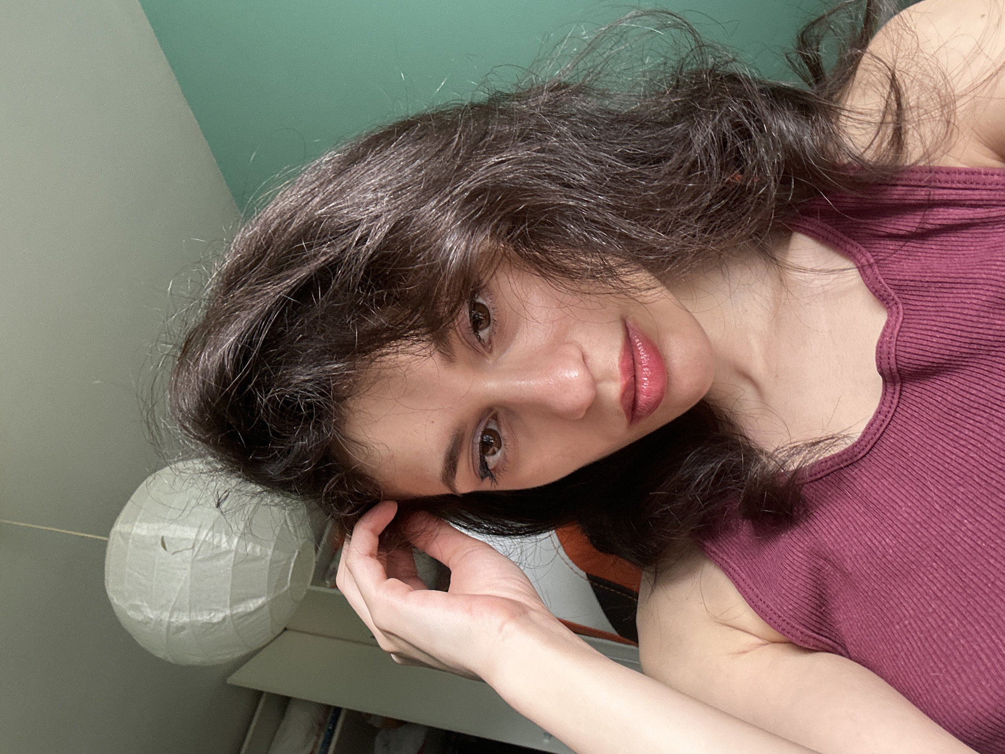 A young woman with long, dark, wavy hair is taking a selfie. She is wearing a dark pink ribbed tank top. Her head is tilted, and her gaze is directed at the camera. Her left hand is touching her hair near her temple. The background shows a wall that is split between a light grey color and a pale green color. To the left of the woman, there is a white paper lantern. The scene appears to be indoors, possibly a bedroom, given the presence of the lantern and what looks like a shelf in the lower right corner. The lighting suggests it is daytime. The woman's expression is neutral and slightly introspective. There is no visible text in the image.