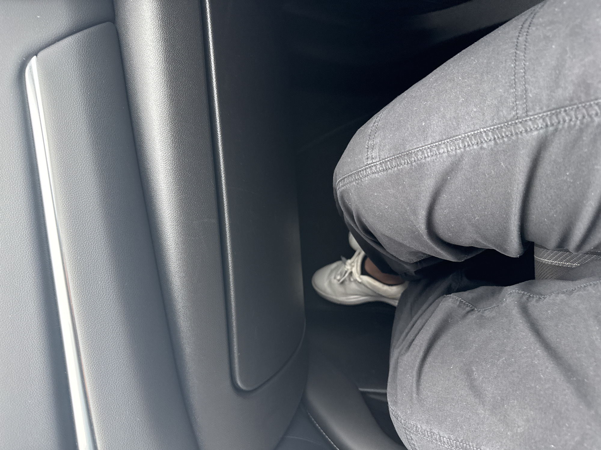 The image shows the interior of a car, with a person's legs and feet visible. The person is wearing black pants and light-colored sneakers. The car's interior is primarily dark grey and black. The location is unknown, but the presence of English writing suggests it is in the United States of America. The city of New Braunfels cannot be confirmed.