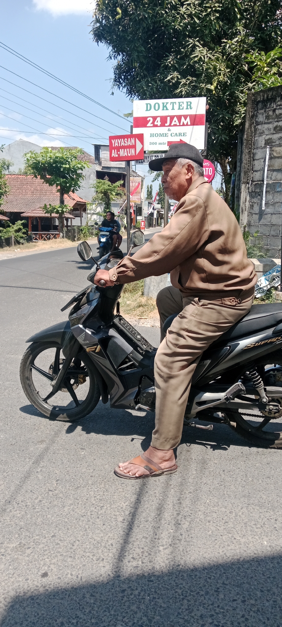 In Maguwoharjo, Indonesia, an elderly man wearing a brown jacket and pants, a black cap, and brown sandals rides a black motorcycle. He is on a paved road with a motorcycle in the foreground and another in the background. A sign in the background reads "DOKTER 24 JAM HOME CARE". Trees and buildings are visible on the left side of the road. The weather appears to be sunny.