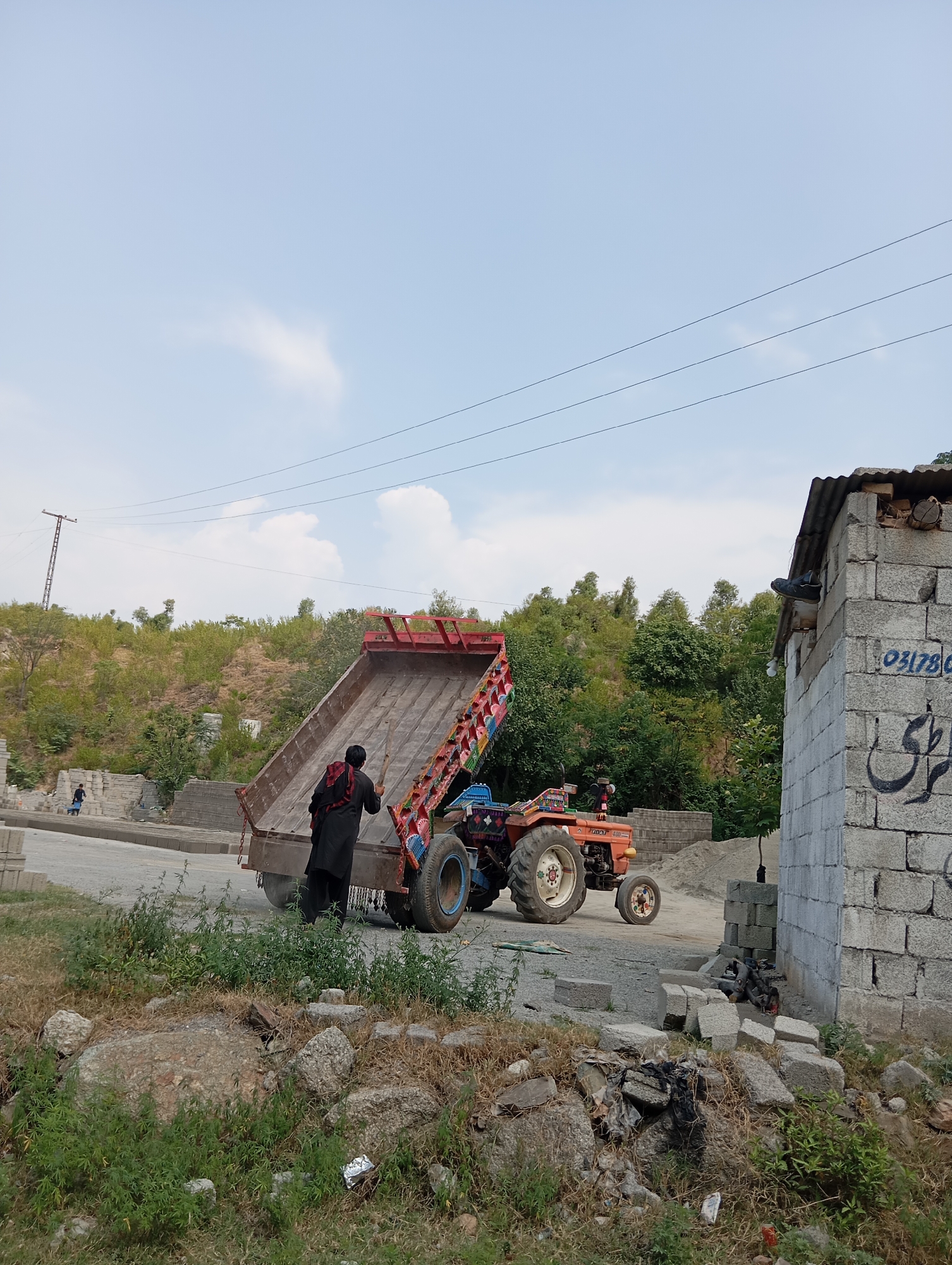 In an outdoor scene, a man dressed in black, with a red scarf around his neck, stands next to a red tractor and its trailer. The trailer is loaded with what appears to be construction materials, possibly bricks or stones. The tractor is orange and blue, and has colorful decorations on its sides. In the background, there are hills covered in vegetation and a blue sky with some clouds. To the right of the scene, there is a structure made of concrete blocks, with a corrugated metal roof. The ground is a mix of dirt, gravel, and some grassy areas with scattered rocks. Another person in blue is visible in the distance to the left, walking near a pile of bricks. Power lines are visible stretched across the sky. The overall impression is that of a construction or transportation site in a rural or semi-rural setting.