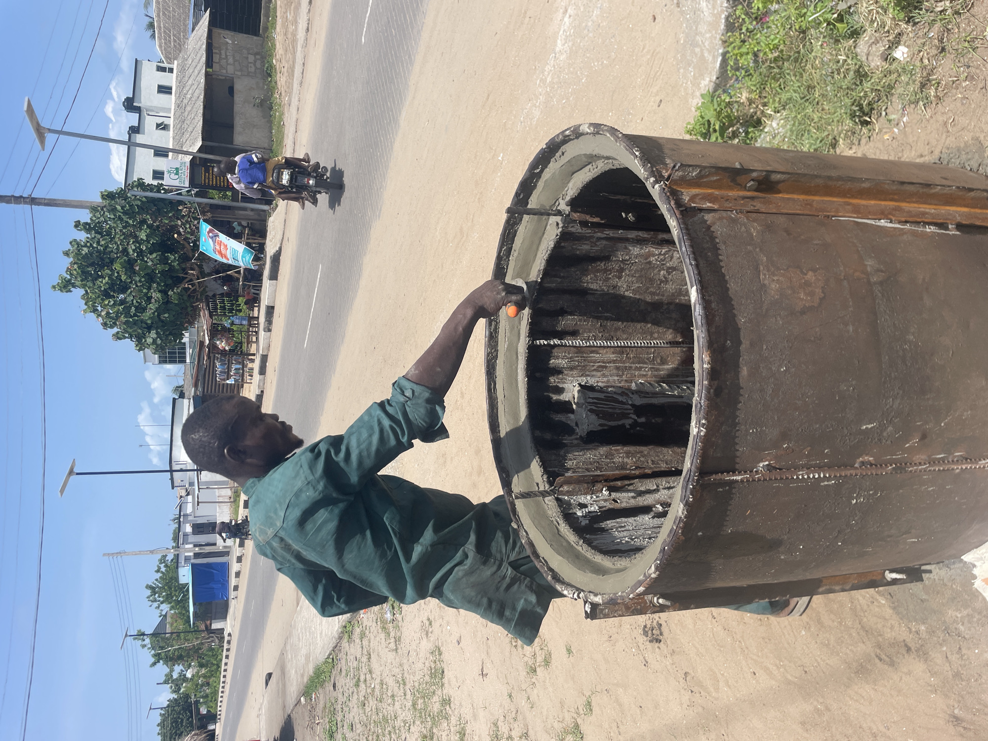 The image captures an outdoor scene in Badagry, Nigeria, under bright daylight with a clear blue sky. The central subject is an adult man, dressed in a green work overall, intently working on a large, rusty, cylindrical metal form or mold. He is applying or smoothing fresh concrete along the inner circumference of the cylinder, with visible rebar (reinforcing bars) inside. He holds a tool with an orange handle, likely a trowel, for this task.
The activity suggests the manufacturing of concrete pipes, culverts, or similar precast concrete structures. The work takes place on a dusty patch of ground adjacent to a paved road. In the background, a person is seen riding a motorcycle on the road. Across the road, several buildings, some appearing partially constructed or in various states of repair, are visible, along with green vegetation and trees. Modern streetlights, fitted with solar panels, are spaced along the roadside. A blue and white banner, partially obscured, is visible near the road, displaying text that includes "GBL CEMENT." The setting reflects a typical semi-urban or peri-urban environment with ongoing construction or industrial activity.