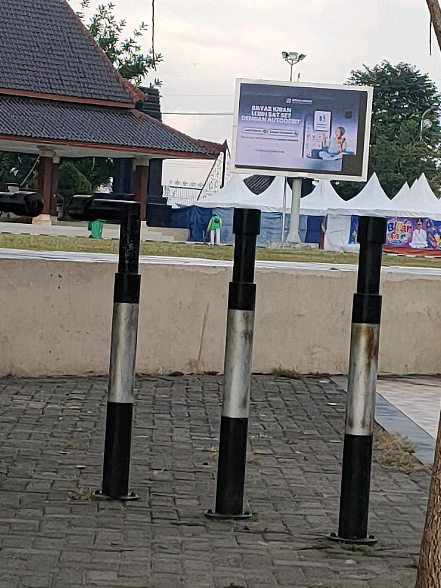 In Kanigoro, Indonesia, a digital billboard displays an advertisement for an automatic bill payment service, featuring text in Indonesian that translates to "Pay electricity bills with AutoDebit." The billboard is mounted on a pole and is positioned behind a row of three metal bollards. In the background, white tents are set up, suggesting an outdoor event or market. A person in a green outfit is visible near the tents, and another person is seen on the billboard advertisement. The scene is set outdoors on a paved area with a stone-like surface.