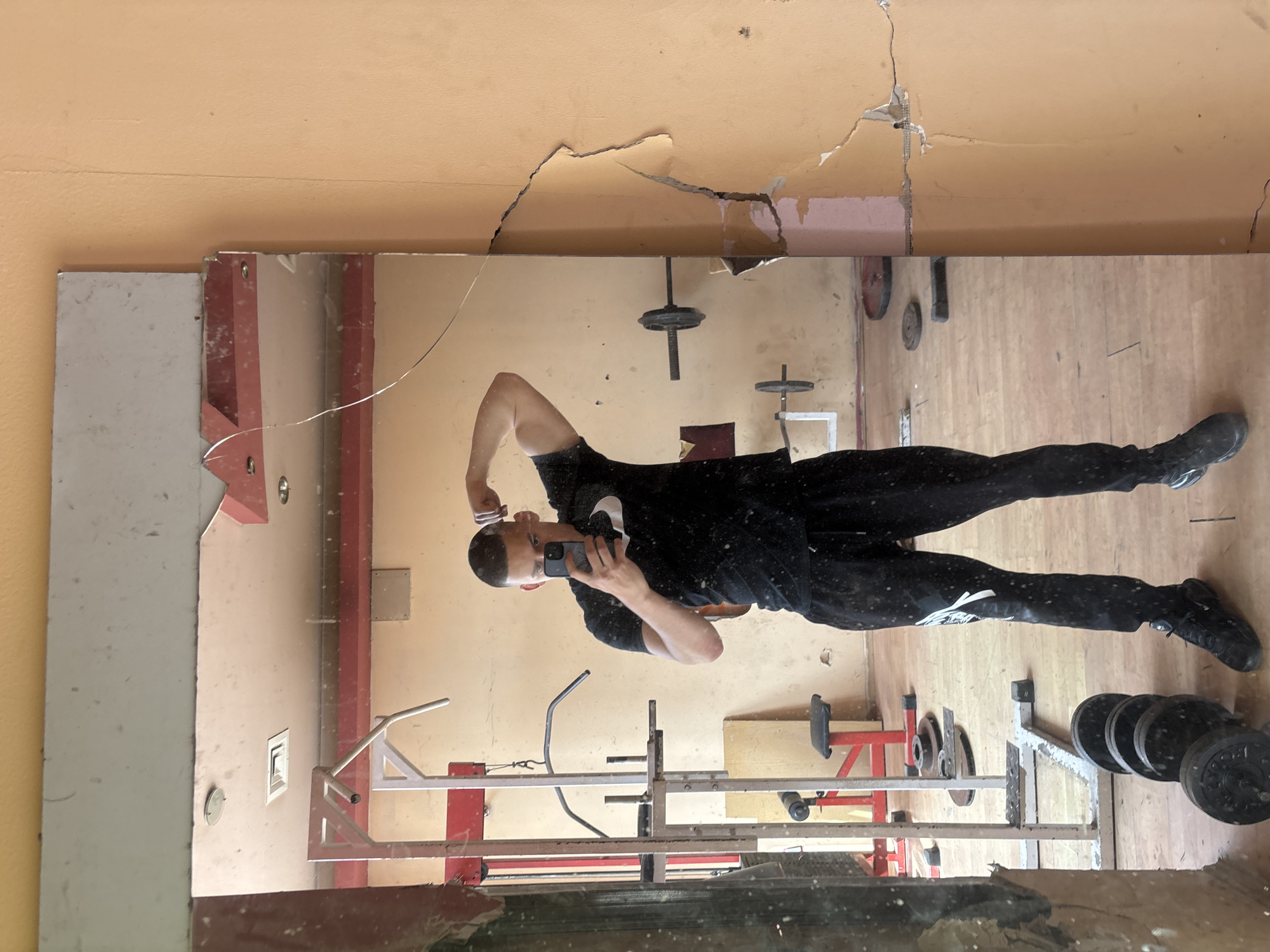 An adult male is captured in a mirror selfie within what appears to be a dilapidated gym or workout area in Goritsa, Bulgaria. The man, seen from the waist up in the reflection, is wearing a black t-shirt with a white logo (possibly a Nike swoosh), black track pants with white accents, and dark sneakers. He is holding a smartphone in his right hand, taking the photo, while flexing his left bicep.
The setting is an indoor space with peach or light orange colored walls that show significant signs of wear, including large cracks and peeling paint, particularly near the top where the wall meets the ceiling. The floor appears to be made of wood. A large, rectangular mirror dominates the scene, reflecting the man and various pieces of gym equipment. The mirror itself is notably damaged, with prominent cracks extending across its surface, especially visible in the upper sections.
Reflected in the mirror, and surrounding the man, are various items of gym equipment. These include several weight plates, some stacked on a stand and others scattered, along with barbell or dumbbell bars. Parts of a red-framed weight bench and a metal squat rack or pull-up bar frame are also visible. The overall impression is one of an old, dusty, and poorly maintained facility, possibly disused. A small rectangular vent is visible on the wall reflected in the mirror. The man's focused expression suggests he is posing for the photograph.