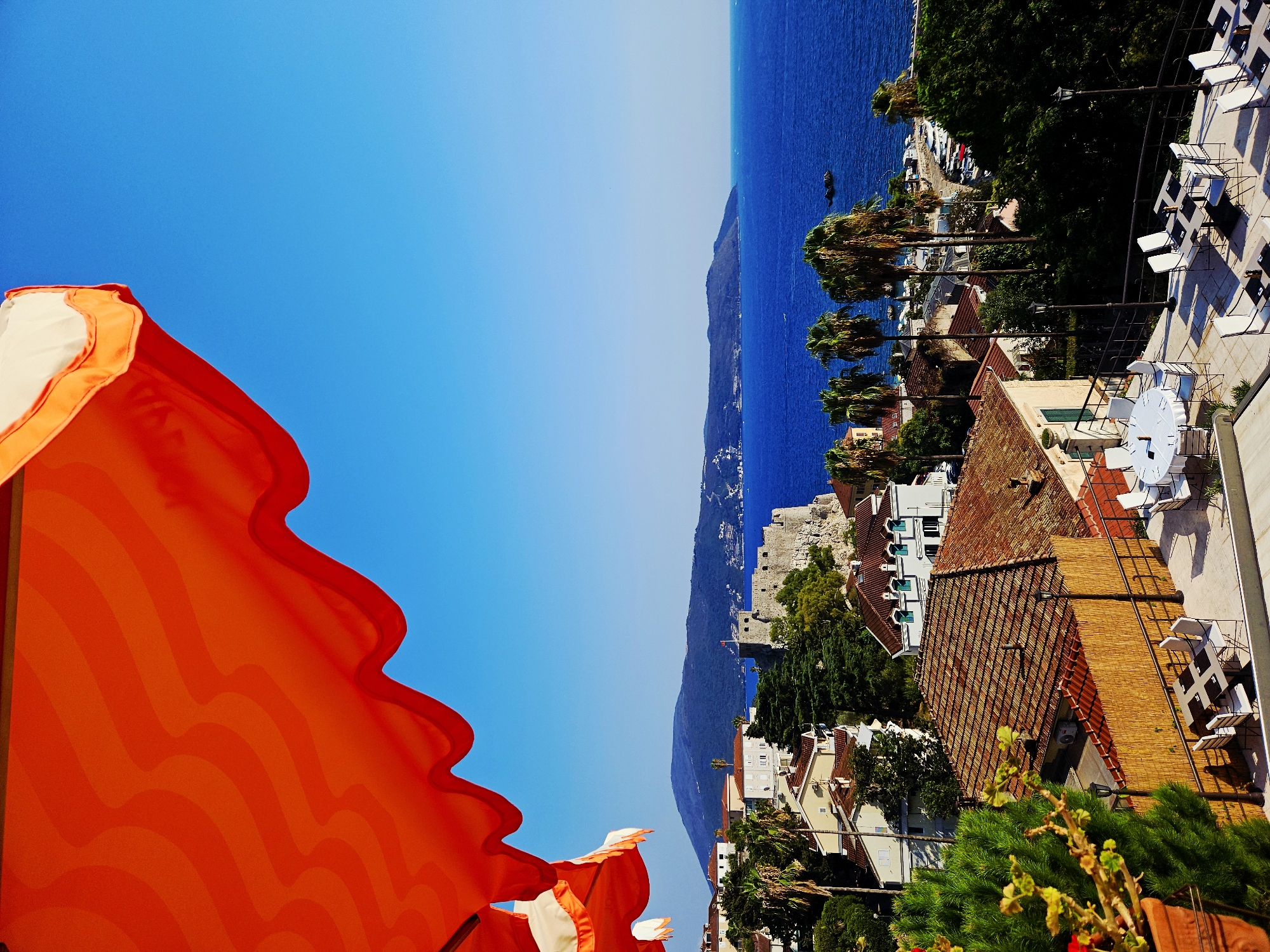 The image depicts a sunny outdoor terrace in Herceg Novi, Montenegro, with a view of the bay. An orange and white awning is visible in the foreground, partially obscuring the clear blue sky. Below the awning, a coastal town with red-tiled roofs and white buildings is nestled against a hillside. A stone fortress with battlements stands prominently on a rocky outcrop overlooking the sea. Lush green trees and palm trees are interspersed among the buildings. The deep blue water of the bay stretches to the horizon, where it meets a hazy mountain range. Several boats are docked along the coastline. The terrace itself features tables and chairs, suggesting a restaurant or cafe setting. The scene is tranquil and bathed in bright sunlight.