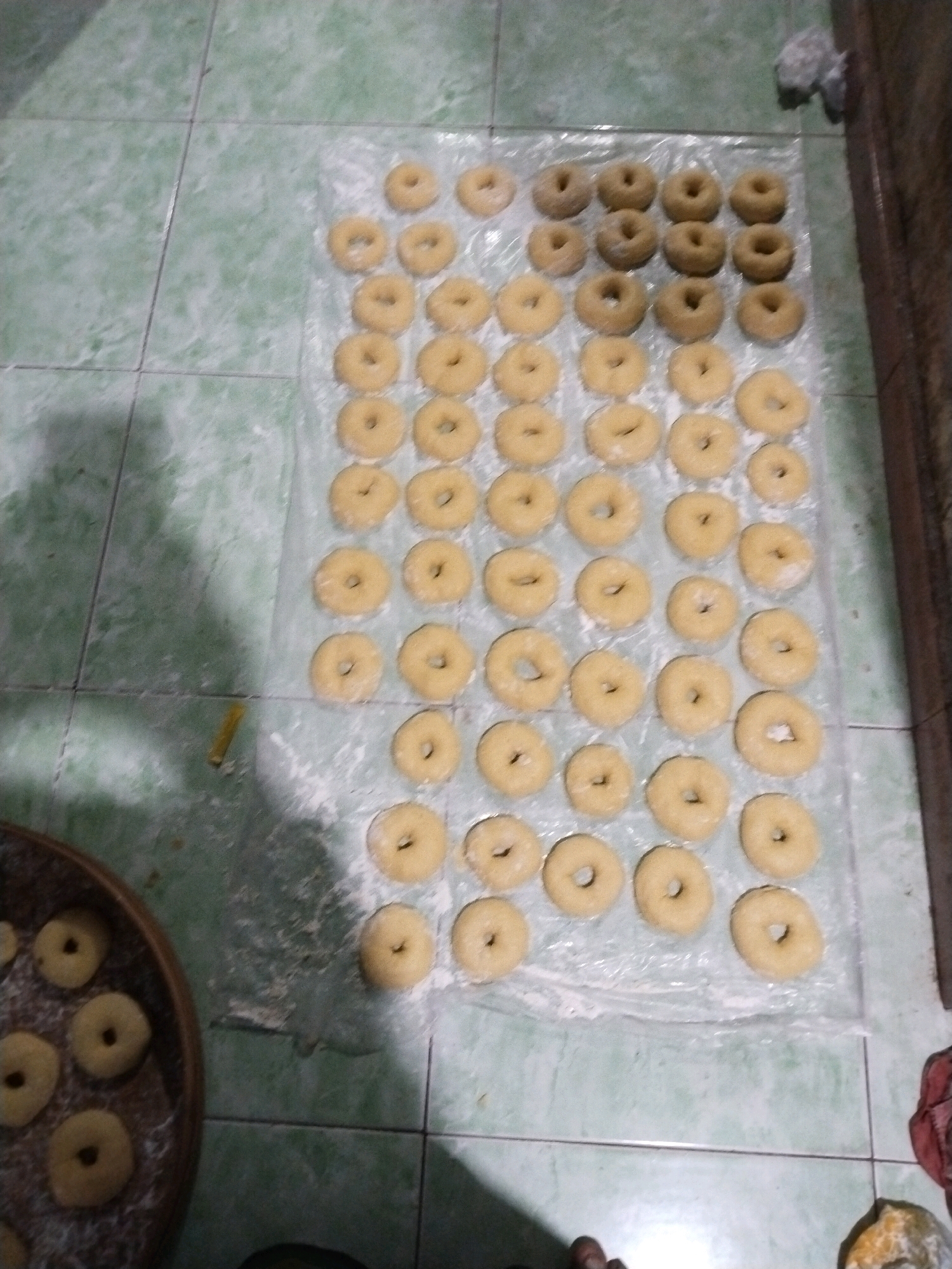 In Tasikmadu, Indonesia, on a floor tiled with green and white squares, a collection of uncooked donuts are laid out on clear plastic sheeting. The donuts are arranged in neat rows and columns, with some appearing slightly larger than others. A woven basket filled with more donuts sits to the left of the main arrangement. The scene suggests food preparation, likely for a local market or personal consumption. The lighting is dim, casting shadows on the tiled floor and obscuring some finer details of the donuts.