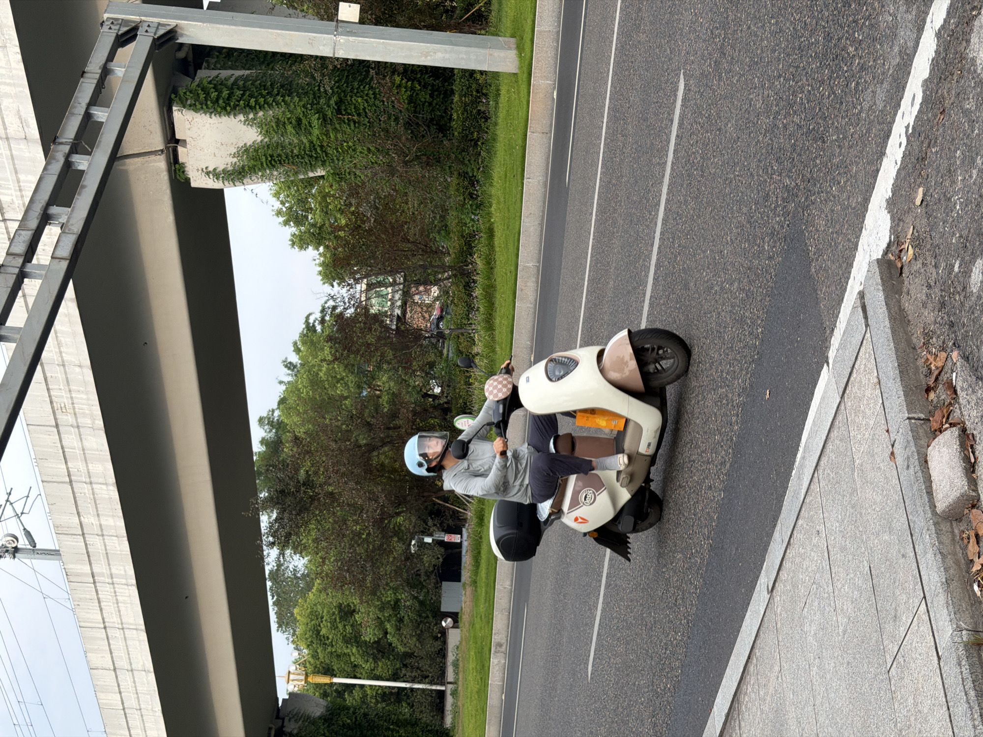 A woman wearing a light blue helmet and a grey hooded jacket rides a cream and tan electric scooter on a paved road in Jiangjin, China. She is wearing dark pants and white slip-on shoes. The scooter has a black storage box on the back and a small orange bag hanging from the front. The scooter has the brand name "Oasis" on its side. The road has white lane markings and is bordered by a grassy median with trees behind it. An overpass with a concrete structure and metal supports is visible above the trees. The ground on the left side of the image is a sidewalk made of grey stone slabs. The lighting suggests it is daytime, likely overcast.