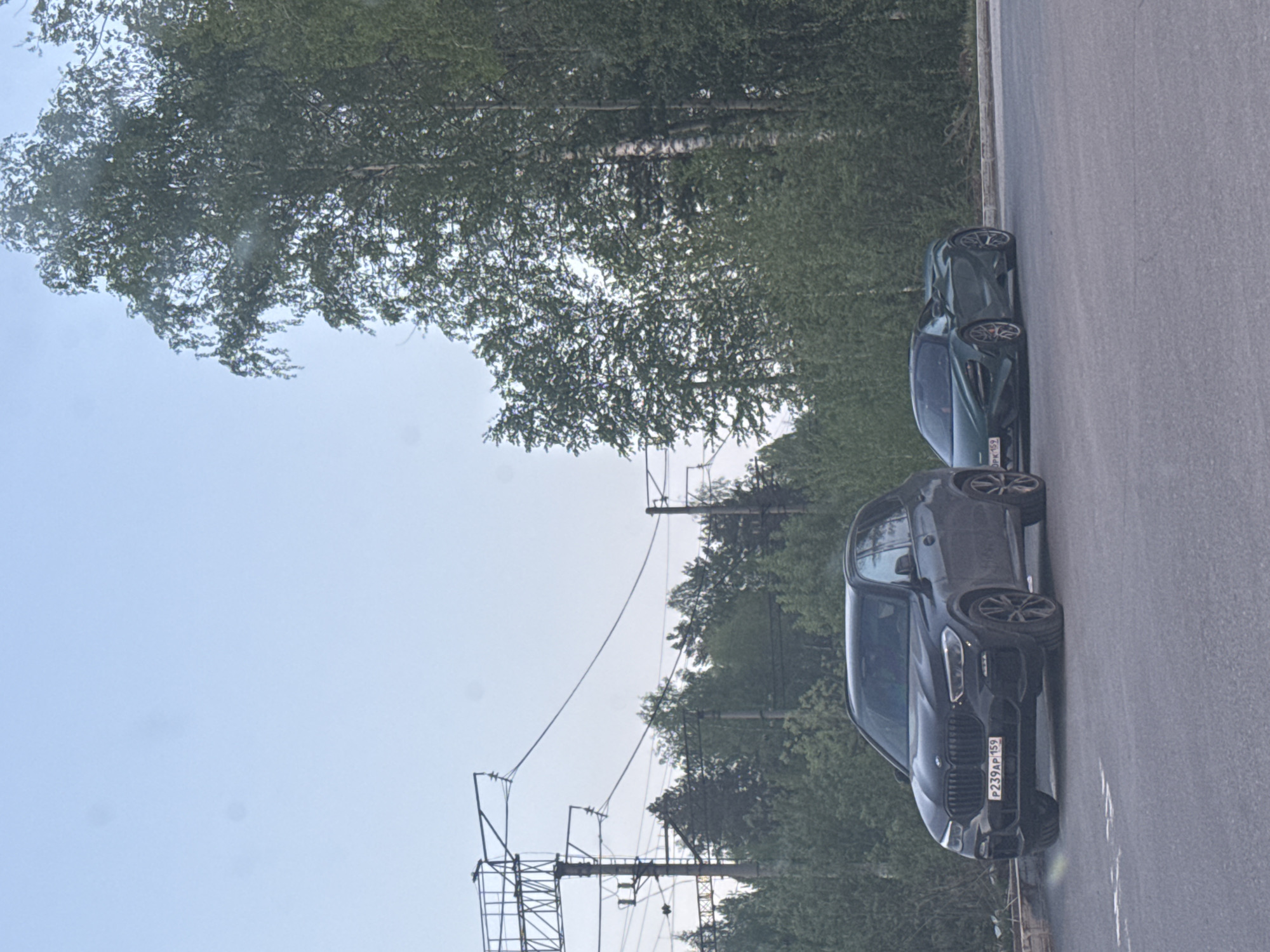 The image shows two cars parked on the side of a road in what appears to be a wooded area. The car closest to the camera is black, with a visible license plate that reads "P239AP 159," which indicates that the location is in the Perm region of Russia. A second car, colored green, is parked behind it. In the background, there are trees and utility poles. The sky is visible above the trees.