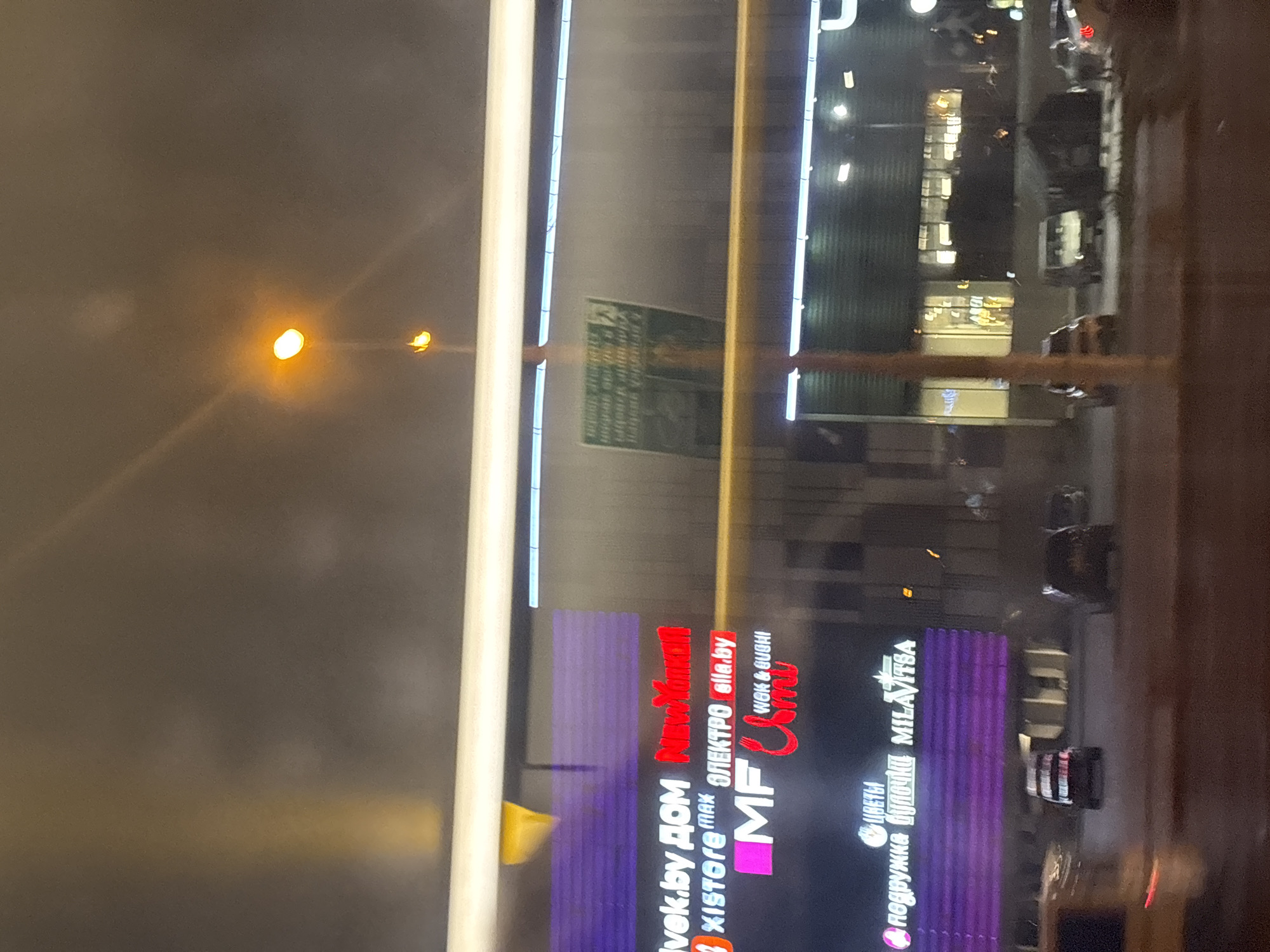 This is a nighttime, slightly motion-blurred exterior shot of a commercial building in Minsk, Belarus. The building features a prominent facade with several illuminated signs. From left to right, visible signs include "ivek.by ДОМ", "xistore MAX", "ЭЛЕКТРО ella.by", and "MF Umi WOK & SUSHI". Below these, there are signs for "ЦВЕТЫ" (Flowers) and "подружка булочки MILAVITSA". The building has a large, illuminated purple section on the lower left. In front of the building, several cars are visible on a street or parking lot, including a dark-colored sedan, a white van, and a dark SUV. The overall environment suggests a commercial district at night, with artificial lighting from the building and streetlights illuminating the scene.