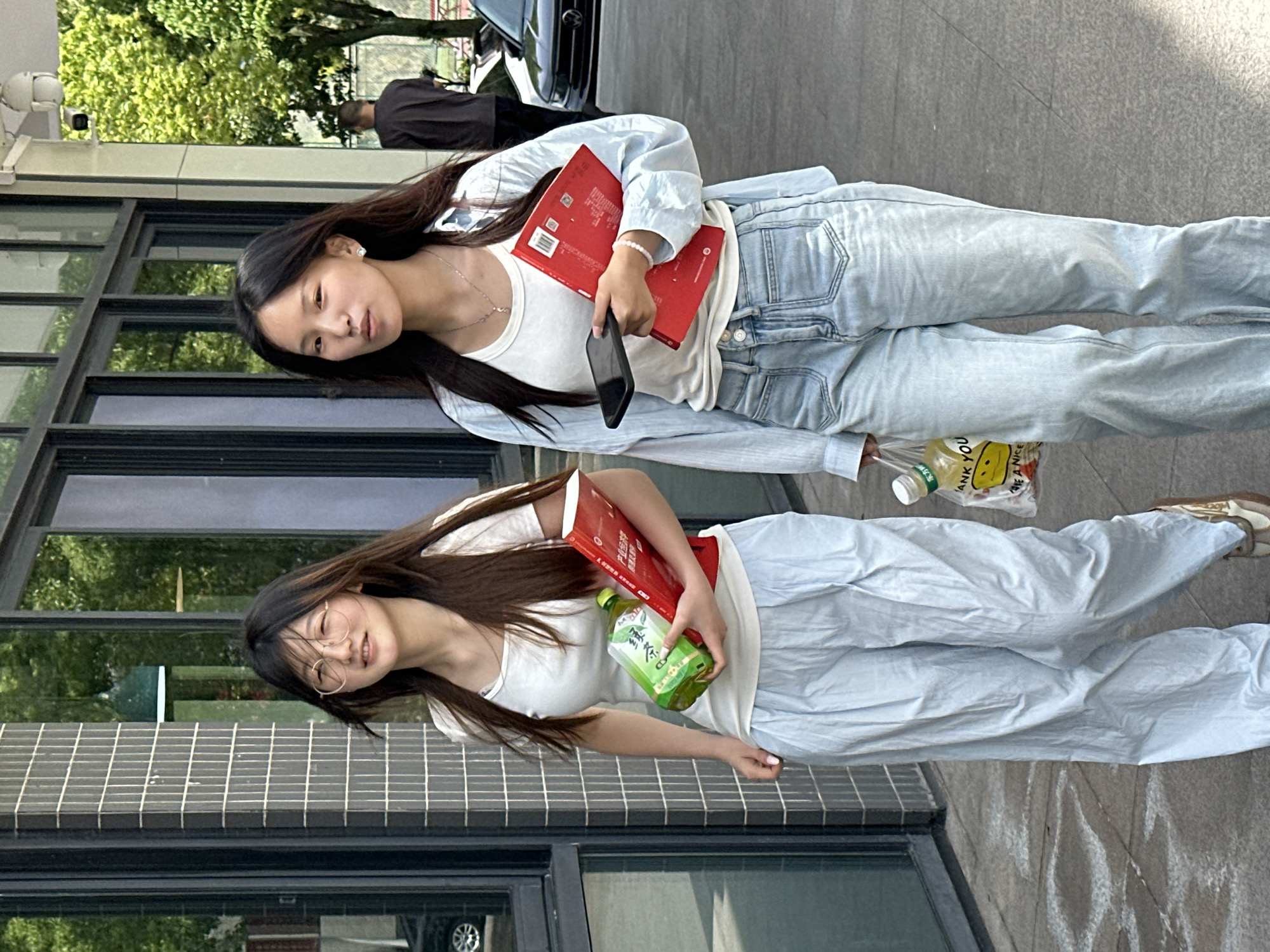 Two young women stand beside a building with windows, holding items. The woman at the top of the image has long dark hair, wears a white top, light denim jeans and jacket, and holds a red book and a black phone. She also carries a plastic bag with a yellow smiley face design. The woman at the bottom has long dark hair, wears a white top, light denim pants, and glasses. She holds a red book and a green bottle. The setting appears to be urban with trees and a street visible in the background. The location is Jiangjin, China. A partial view of a person on a scooter and a black car is also seen.