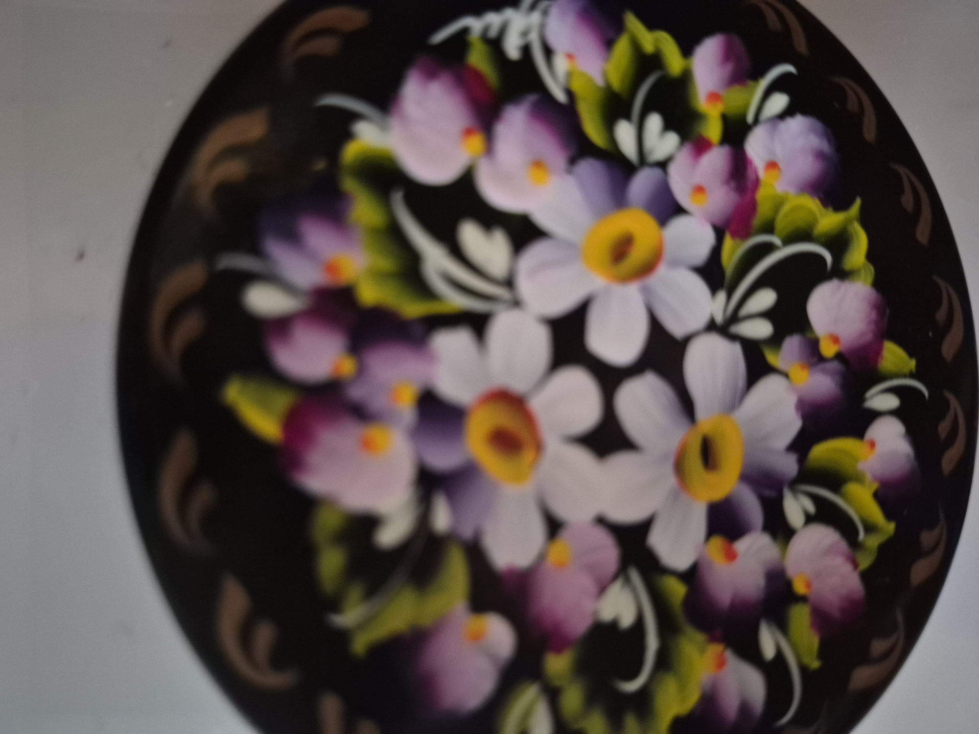 The image shows a close-up of a dark-colored plate or tray decorated with a floral pattern. The flowers are painted in shades of white, purple, and pink, with yellow centers. Green leaves and white accents are also visible. The background is blurred and appears to be a light gray surface, possibly a table or wall. There is no discernible information in the image that would indicate its location in the city of Vanadzor, Armenia.