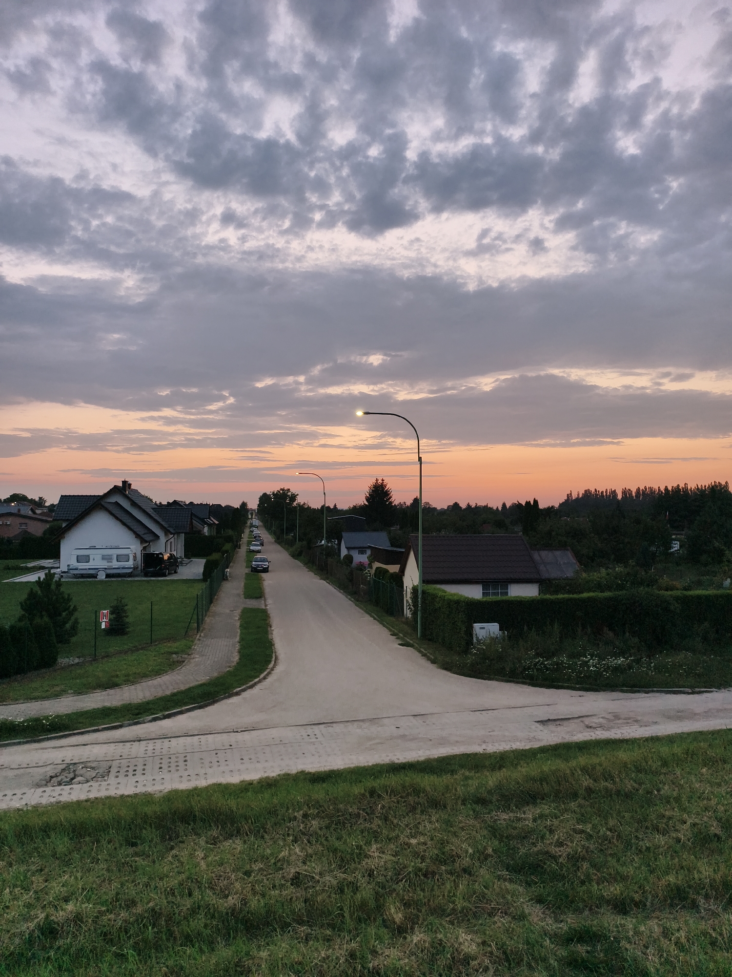 The image shows a residential street in Tczew, Poland, during what appears to be sunset. The sky is filled with dramatic, cloudy formations in shades of gray and pink, with a warm orange glow near the horizon. A paved road runs from the foreground into the distance, lined with houses and lush green trees. Several cars are parked along the street. In the foreground, a grassy embankment overlooks the road, and to the left, there is a section of textured concrete. Streetlights are visible along the road, one of which is illuminated. The scene depicts a quiet evening in a suburban area.