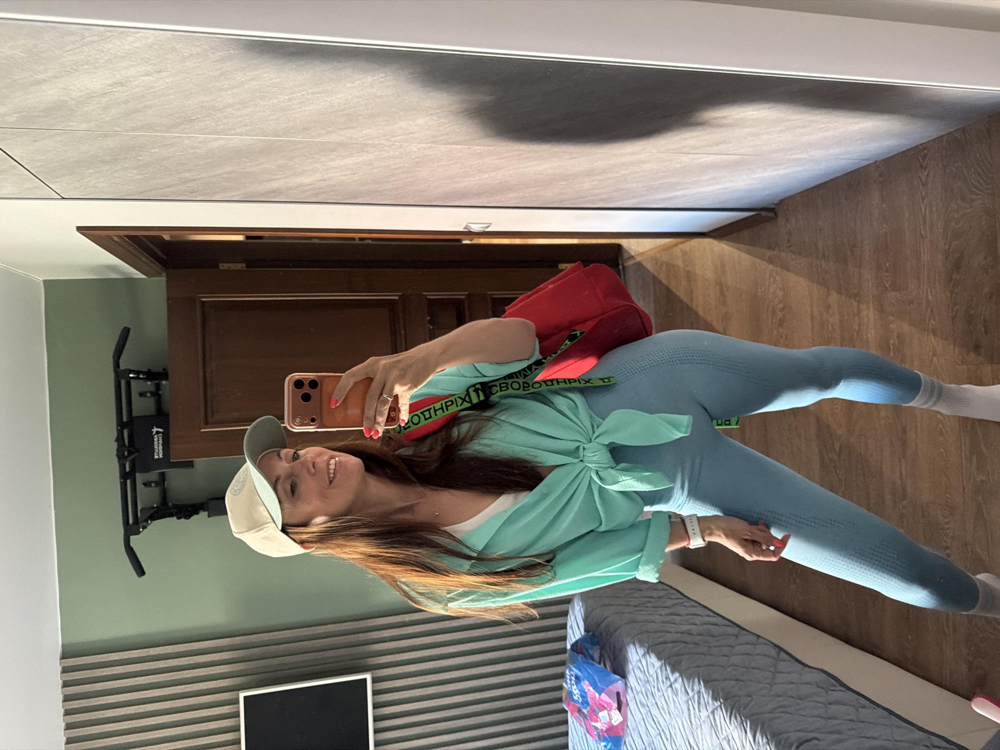 A woman stands in a room, taking a mirror selfie. She is wearing a light blue tie-front shirt, a white sports bra, light blue leggings, a beige baseball cap, and grey socks with white stripes. A red bag with a black and red strap hangs over her shoulder. She is holding an orange smartphone in her right hand. Behind her, a bed with a grey quilted cover is visible, and to her right, a large grey wardrobe stands. A pull-up bar is mounted on the wall above a wooden door. The walls are painted a light green color, and the floor is made of wood-look laminate. The lighting suggests it is daytime, with sunlight casting shadows on the floor and wardrobe. This scene is set in a residential interior, not specifically identifiable as being in Birobidzhan, Russia.