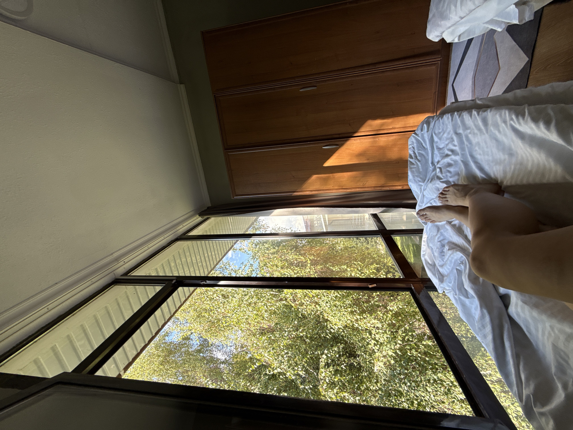 In the city of Sary-Oy, Kyrgyzstan, a first-person perspective shot captures a bedroom scene illuminated by sunlight streaming through a large, multi-pane window. The window offers a view of lush green trees and a blue sky. In the foreground, a person's legs and feet are visible, resting on a white duvet-covered bed. To the right of the bed, a tall, wooden wardrobe with three doors stands against a dark green wall. Sunlight casts distinct rectangular shadows across the wardrobe doors and the floor, where a geometric rug is laid out. The ceiling is white, with visible molding along the top of the walls.