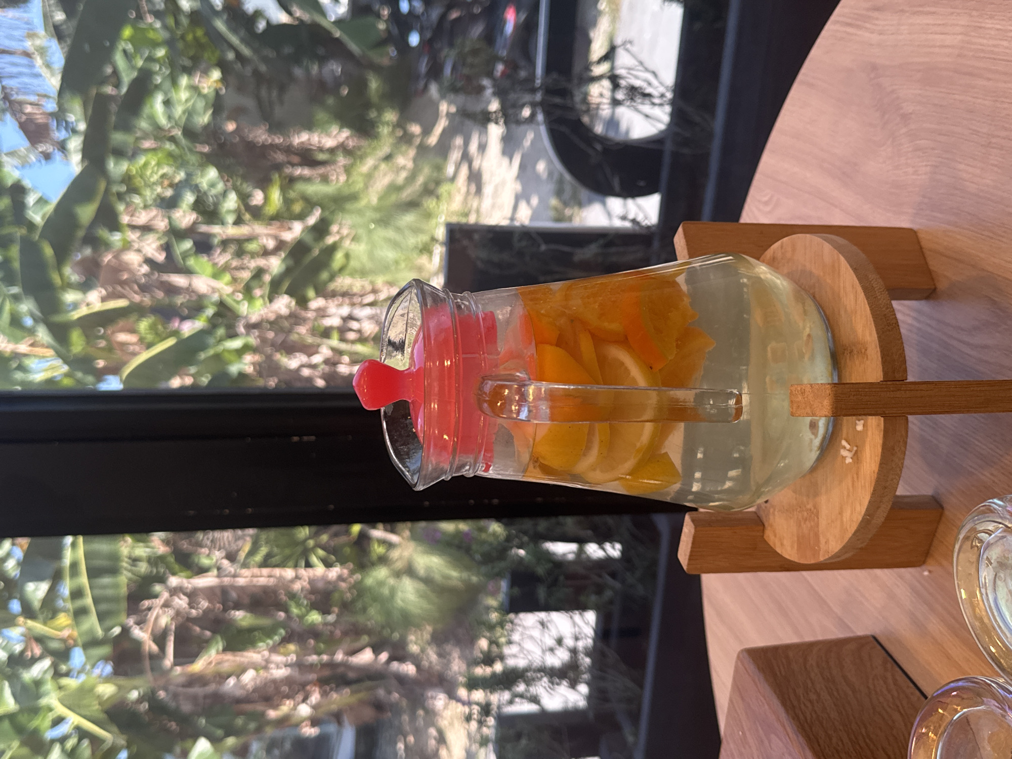 A pitcher of citrus-infused water sits on a wooden stand on a table. The pitcher has a red lid and is filled with water and slices of oranges and lemons. In the background, through a window, is a blurred view of palm trees and foliage, suggesting an outdoor or tropical setting. The scene appears to be in Kuta Selatan, Indonesia, likely at a cafe or restaurant given the presentation and the presence of glassware in the foreground.
