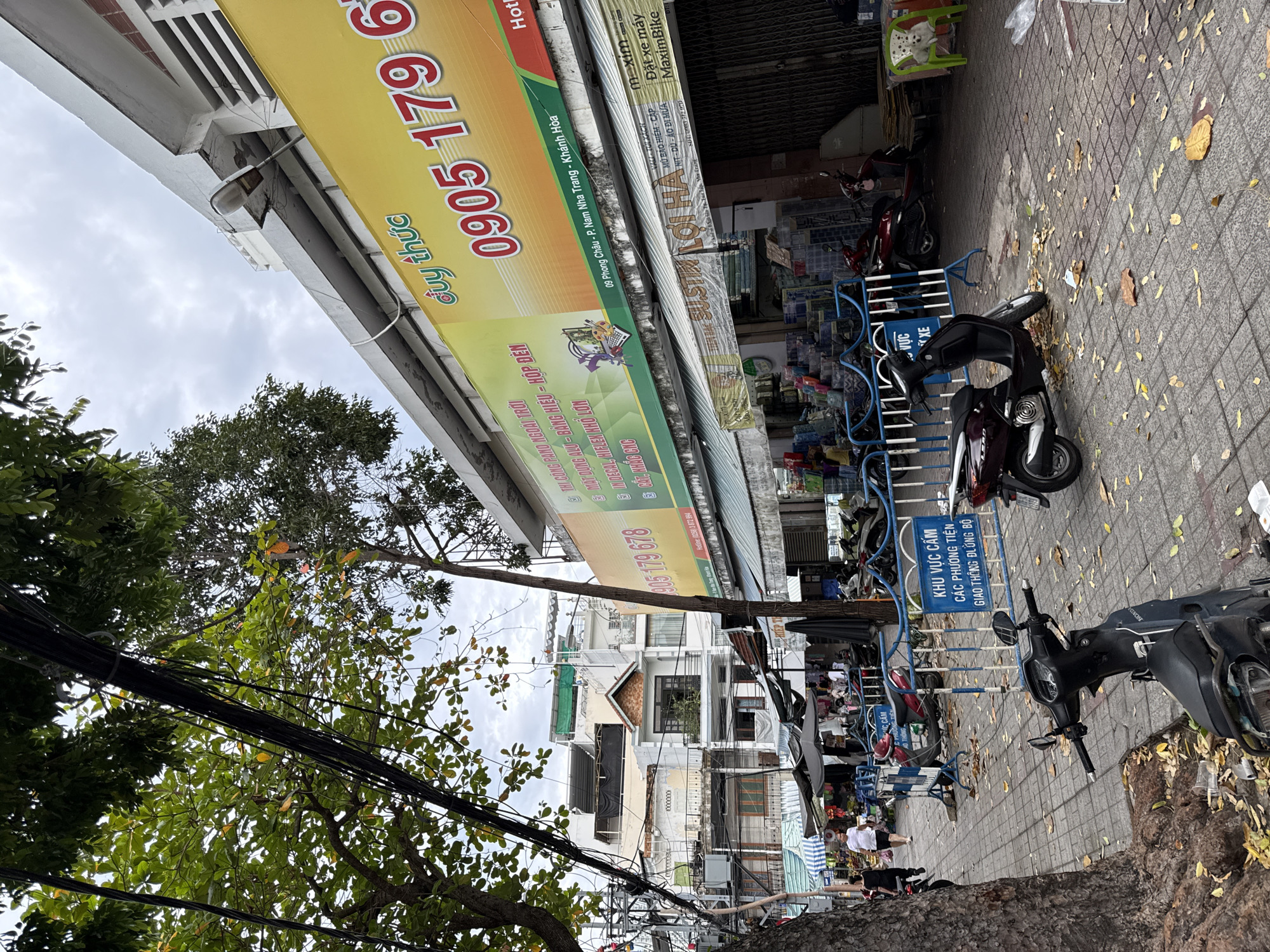 The image shows a street scene in Nha Trang, Vietnam. A yellow banner with red lettering advertising "DUY THỨC" and a phone number "0905 179 678" is visible on the side of a building. Below the banner, a row of shops with awnings is set up along the sidewalk. In the foreground, several motorcycles are parked on the paved sidewalk, some of which are behind a blue metal barrier. A sign on the barrier reads "KHU VỰC CẤM CÁC PHƯƠNG TIỆN GIAO THÔNG ĐƯỜNG BỘ", indicating a restricted area for vehicles. Fallen leaves are scattered on the sidewalk. A few people are visible in the background, walking along the street and near the shops. The sky is overcast.