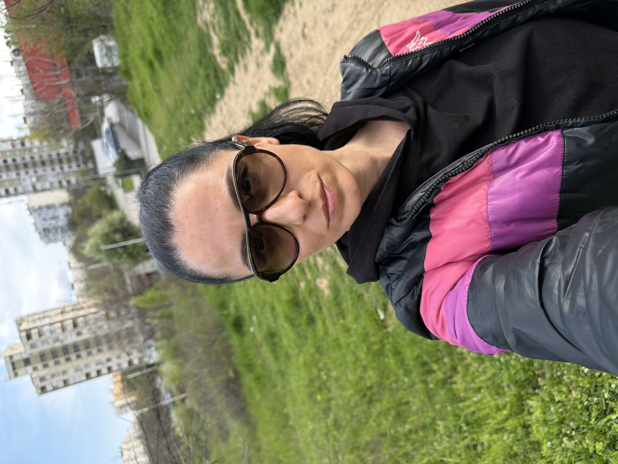 A woman with dark hair pulled back and wearing sunglasses poses for a selfie outdoors. She is wearing a black hooded sweatshirt underneath a black jacket with pink and purple panels on the shoulders. The background shows an urban environment with grass, trees, and buildings. Based on visual landmarks and urban layout, the location appears to be in the city of Pleven, Bulgaria. Specifically, the buildings are reminiscent of the architecture in the areas near the Kaylaka park.
