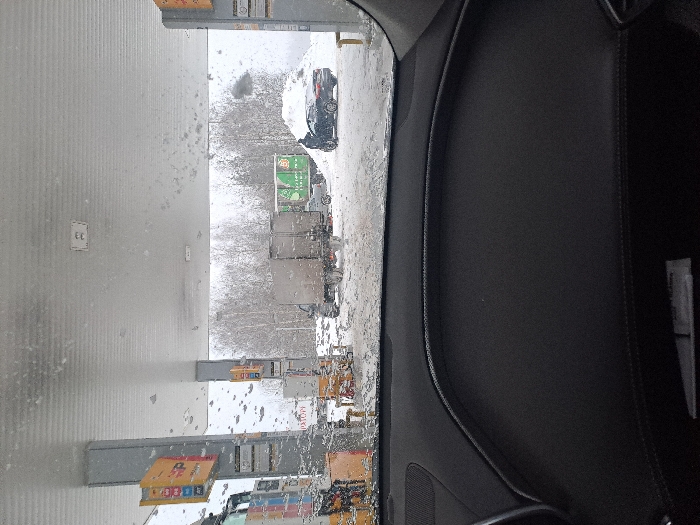 A snowy scene is viewed from inside a vehicle. Several trucks are lined up in a parking lot, visible through the windshield, which is covered in droplets. A grey building stands to the left of the trucks. The interior of the car is visible on the right, including a dark dashboard.