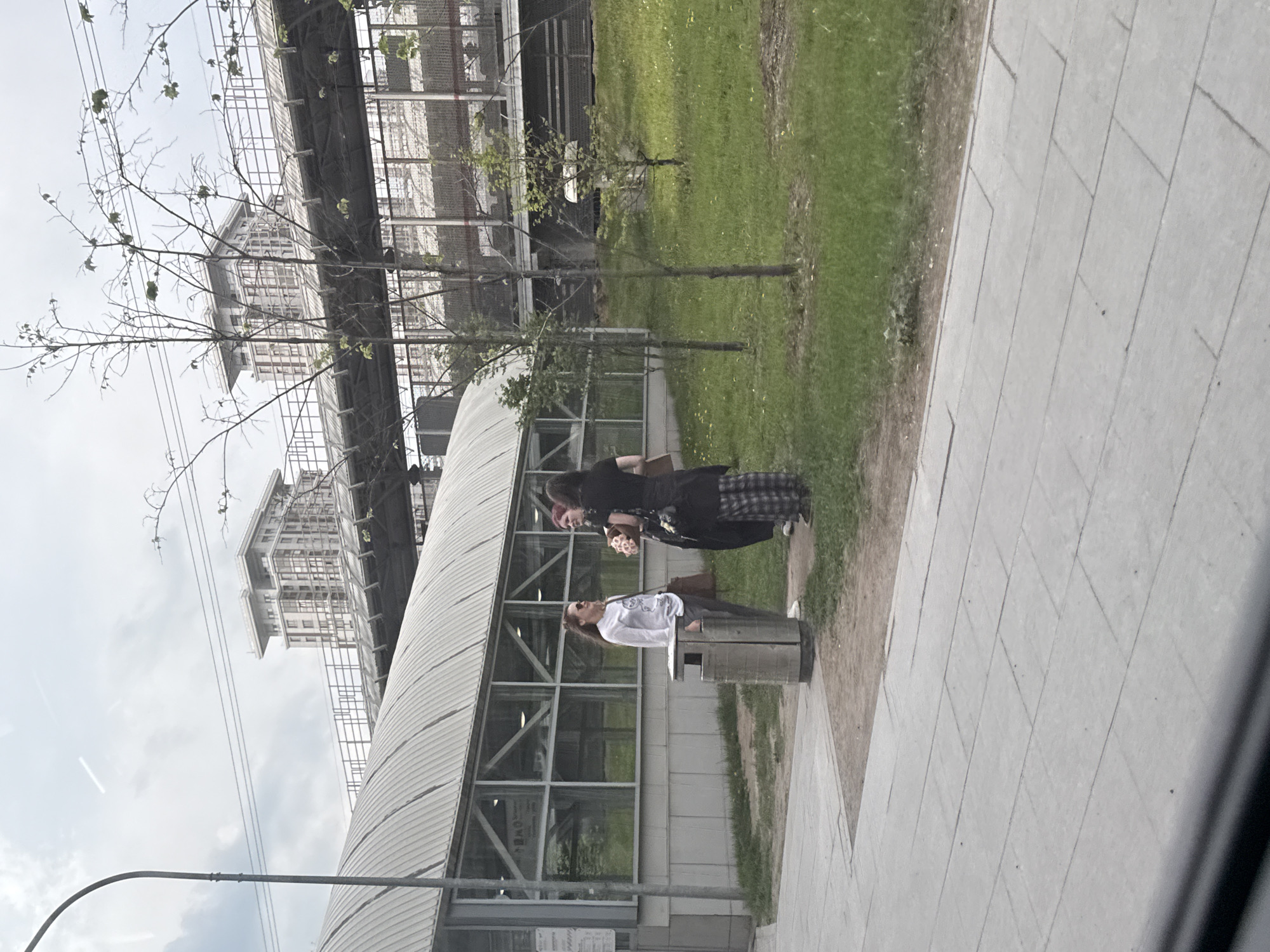 The image depicts a scene in Moscow, Russia. In the foreground, two people stand on a paved walkway next to a metal trash receptacle. One person, wearing a white shirt and sunglasses, leans against the receptacle. The other person, dressed in black with a plaid skirt, stands beside them, holding an object. Behind them is a modern structure with a metal roof and glass windows. In the background, a large building under construction is visible, supported by metal scaffolding. A small patch of green grass is also present. A lamppost is seen in the lower left of the frame.