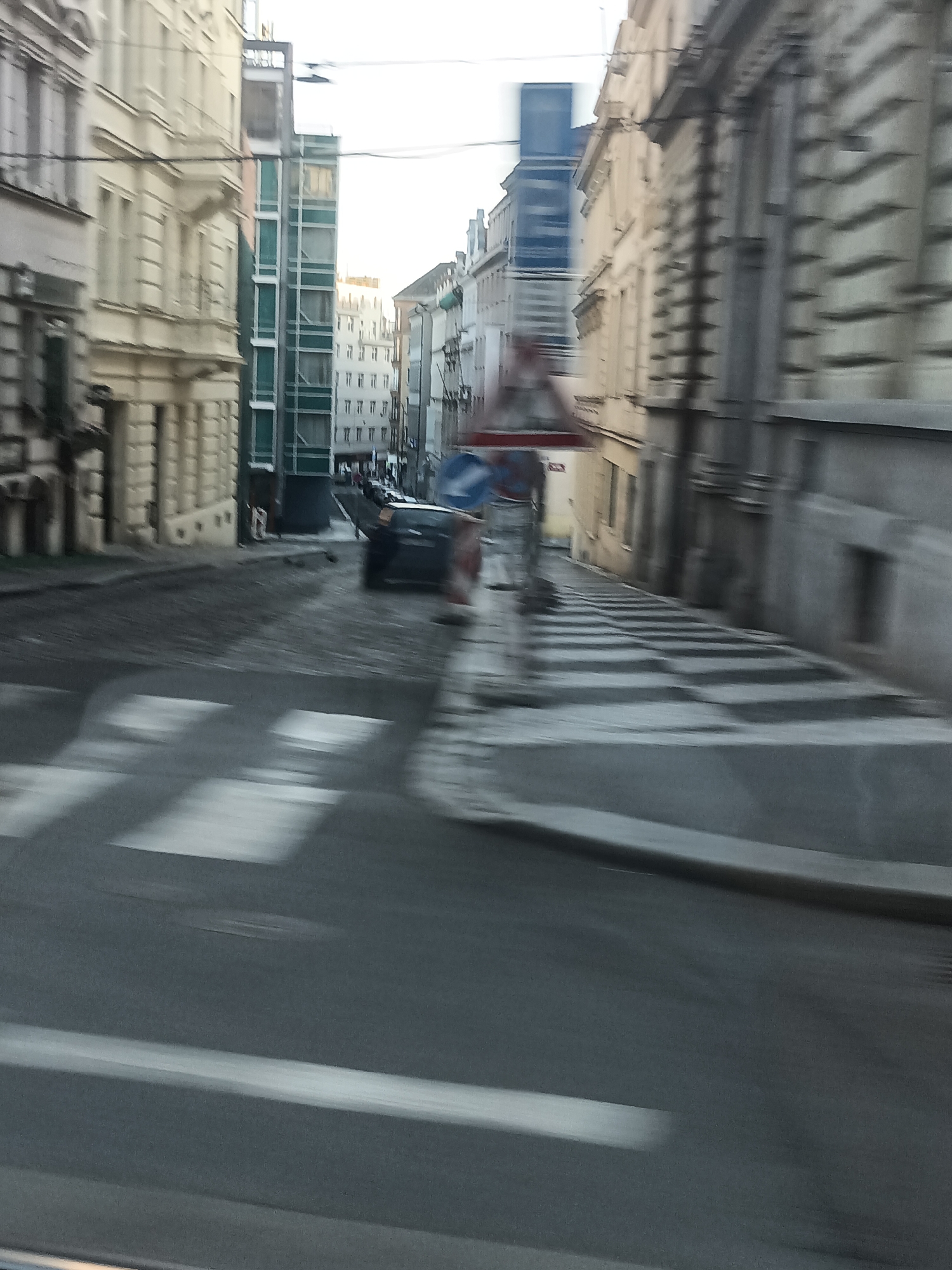 The image depicts a city street scene in Prague, Czechia. The perspective is from a moving vehicle, indicated by the motion blur. In the foreground, a pedestrian crossing with white stripes is visible on the asphalt road. Buildings line both sides of the street, with a mix of architectural styles. On the left, a light-colored building with multiple windows and decorative elements is prominent. On the right, a darker, more modern building with large windows is visible. Further down the street, a blue traffic sign is present, and a dark-colored car is moving. The overall impression is a dynamic urban environment with a sense of movement.