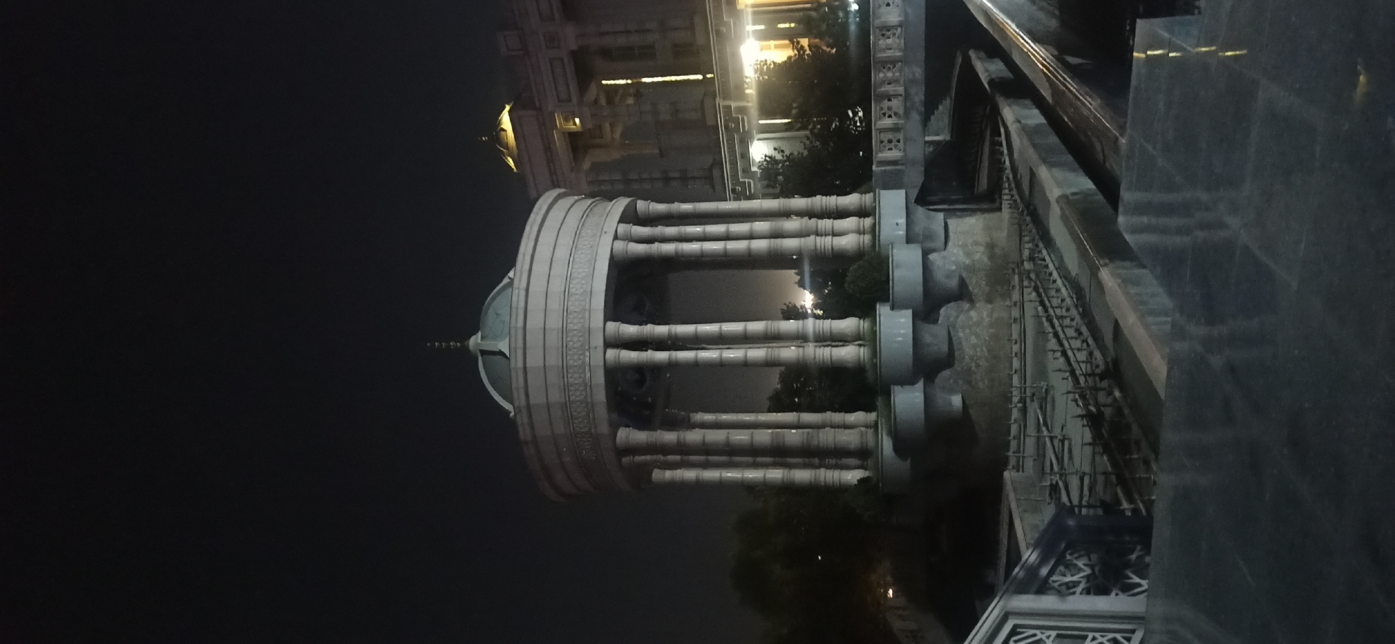 The image shows a nighttime view of the Rudaki Park in Dushanbe, Tajikistan. The central subject is a domed rotunda supported by fluted columns, with an ornate frieze running around the upper drum. The structure is illuminated and appears to be made of light-colored stone. Behind the rotunda, a larger, modern building with multiple illuminated windows and a golden dome is visible. In the foreground, a fountain or water feature with various channels and structures is present, some of which appear to be under construction or maintenance. Trees are visible to the left of the rotunda, and the sky is dark. The overall impression is a well-lit public space at night.