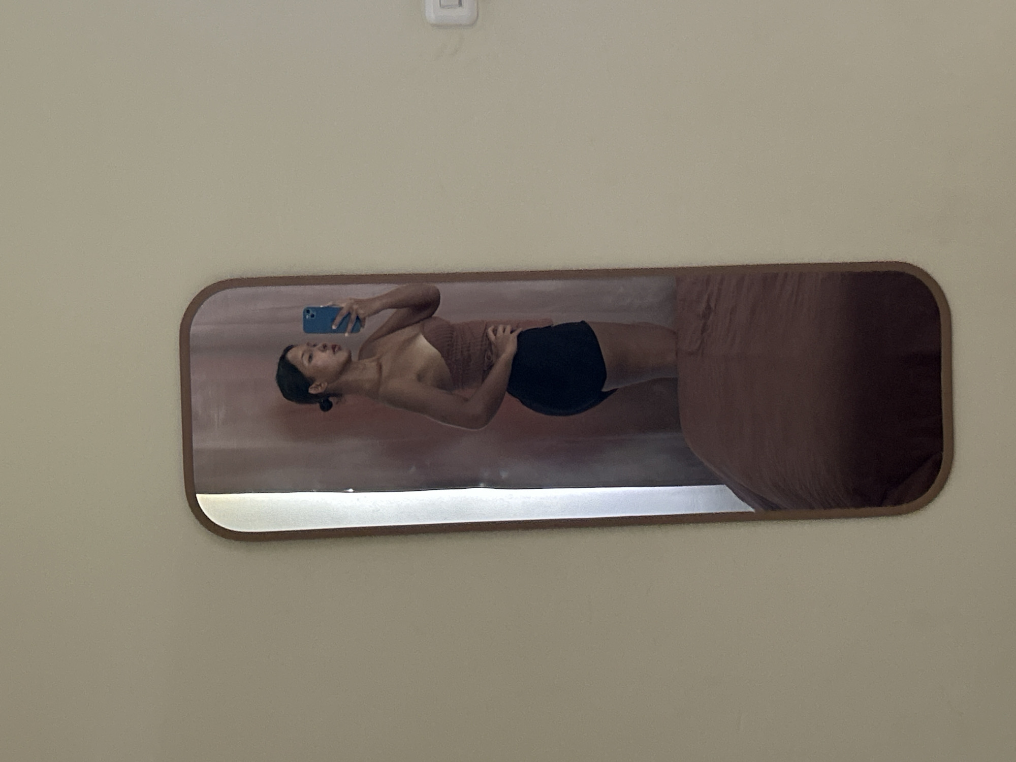 In Kuta, Indonesia, a young woman with dark hair pulled back in a bun stands in front of a full-length mirror, taking a selfie with a blue smartphone. She is wearing a strapless, ruched brown top and black shorts. The mirror is framed in a brown wood, and behind her, a bed with a dark reddish-brown comforter is partially visible. The wall behind the mirror is a light beige color.