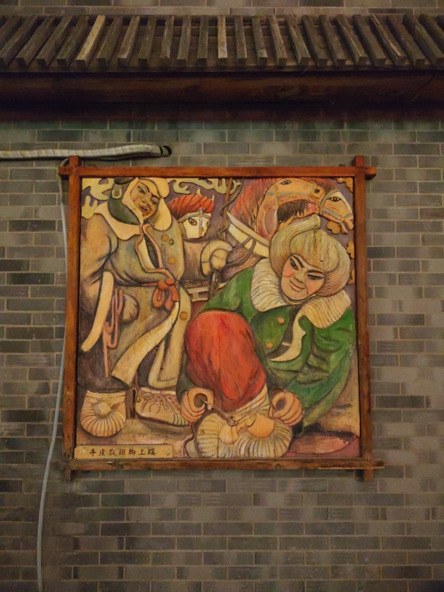The image shows a framed wooden carving depicting several figures and horses. The carving is mounted on a brick wall. At the top of the image, a wooden roof structure is visible, with overlapping wooden slats. Below this, a white cable runs horizontally across the brick wall. The carving itself is colorful, with figures in traditional clothing. In the foreground, a figure in a green garment with a fur collar smiles, with a red-capped figure leaning towards them. Behind them, other figures and horses are depicted. At the bottom of the carving, there is Chinese text. The brick wall appears to be dark grey and is laid in a standard running bond pattern. The overall scene suggests an indoor or covered outdoor display, likely a cultural or artistic installation.