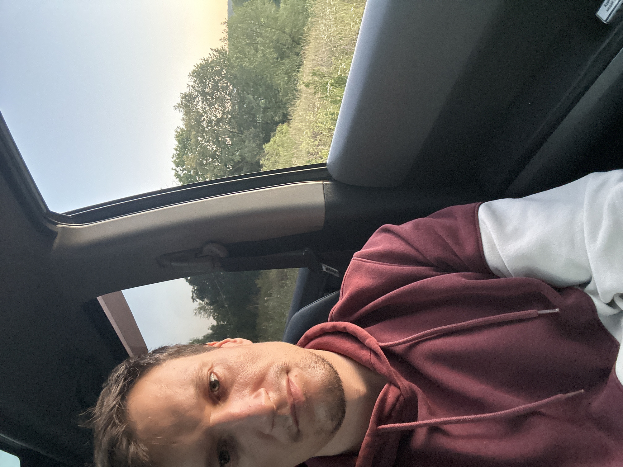 A close-up, selfie-style shot captures a light-skinned male with brown hair and a slight beard looking directly at the camera. He is wearing a maroon hoodie with white sleeves. The man is seated inside a vehicle, with the interior of the car visible, including the door panel and a seatbelt. Outside the car window, a rural landscape unfolds, featuring trees and tall grass under a clear sky, suggesting an outdoor setting likely on the outskirts of Makiv, Ukraine. The lighting indicates it may be late afternoon or early evening.