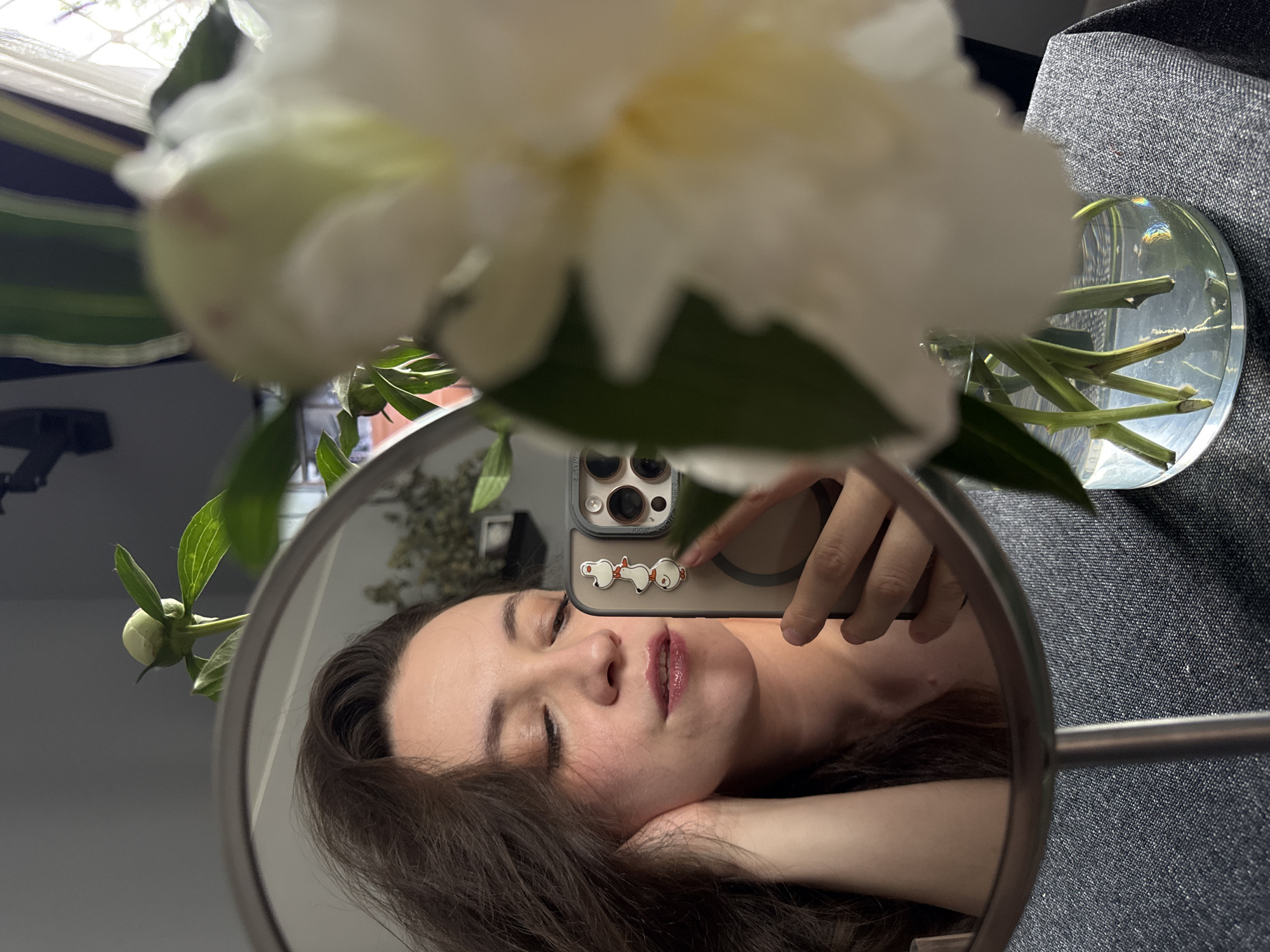 In this close-up mirror selfie, a young woman with long, dark brown hair is captured from the shoulders up. She is looking down and to her left, with her left hand resting on her cheek and her arm bent at the elbow. Her eyes are closed, and her lips are slightly parted, with a glossy, pinkish lipstick. She is wearing a light-colored top.
Her reflection is visible in a round, silver-toned mirror. She is holding a smartphone in her right hand, angled towards the mirror to take the photo. The phone case is light beige and adorned with a vertical stack of three small, white cartoon ducks. The phone's camera lenses are prominent.
In the foreground, out of focus, is a large, white peony bloom in a clear glass vase filled with water. The flower's petals are soft and slightly translucent. Several green stems and leaves are visible submerged in the water. The vase sits on a dark gray, textured surface, possibly a tablecloth or fabric.
In the background, reflected in the mirror, is a room with a gray wall. A green plant with large leaves is visible above the woman's head, to the left, with a closed bud on one of its stems. To the right, a window with sheer white curtains and a diamond pattern is visible. The overall lighting suggests an indoor setting, possibly near a window. There are no discernible elements that would place this scene specifically in the city of Chaikovsky, Russia.