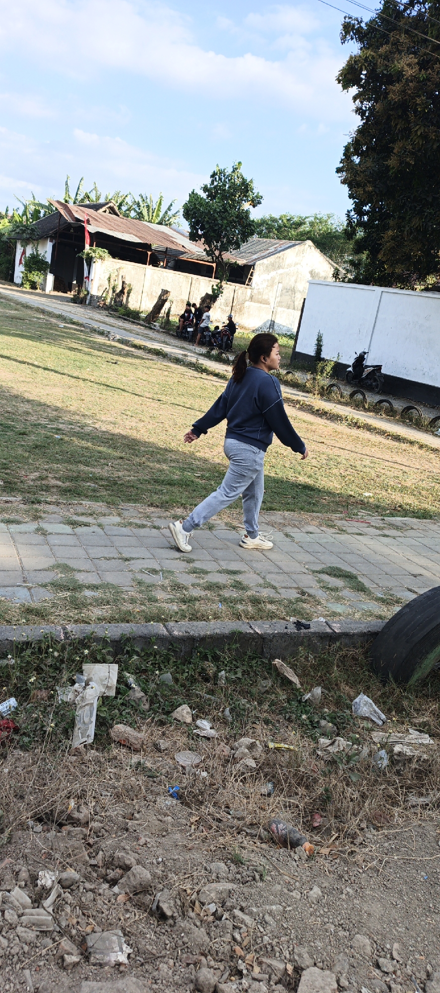 In Kasiyan Timur, Indonesia, a woman with dark hair pulled back in a ponytail is captured in motion, walking on a paved walkway. She is wearing a dark blue long-sleeved sweatshirt and light gray sweatpants, paired with white sneakers. Her head is turned to her right, and she appears to be looking away from the camera. The walkway is bordered by a grassy area on the left and a dirt and debris-filled embankment on the right, which includes a large tire.
In the background, a cluster of simple structures with corrugated metal roofs and concrete walls suggests a residential or community area. Several motorcycles are parked near these buildings, and some individuals are seated on them, with one person standing. Beyond the structures, lush green trees and vegetation are visible against a bright blue sky dotted with white clouds. The overall scene depicts an outdoor setting with a mix of activity and natural elements.
