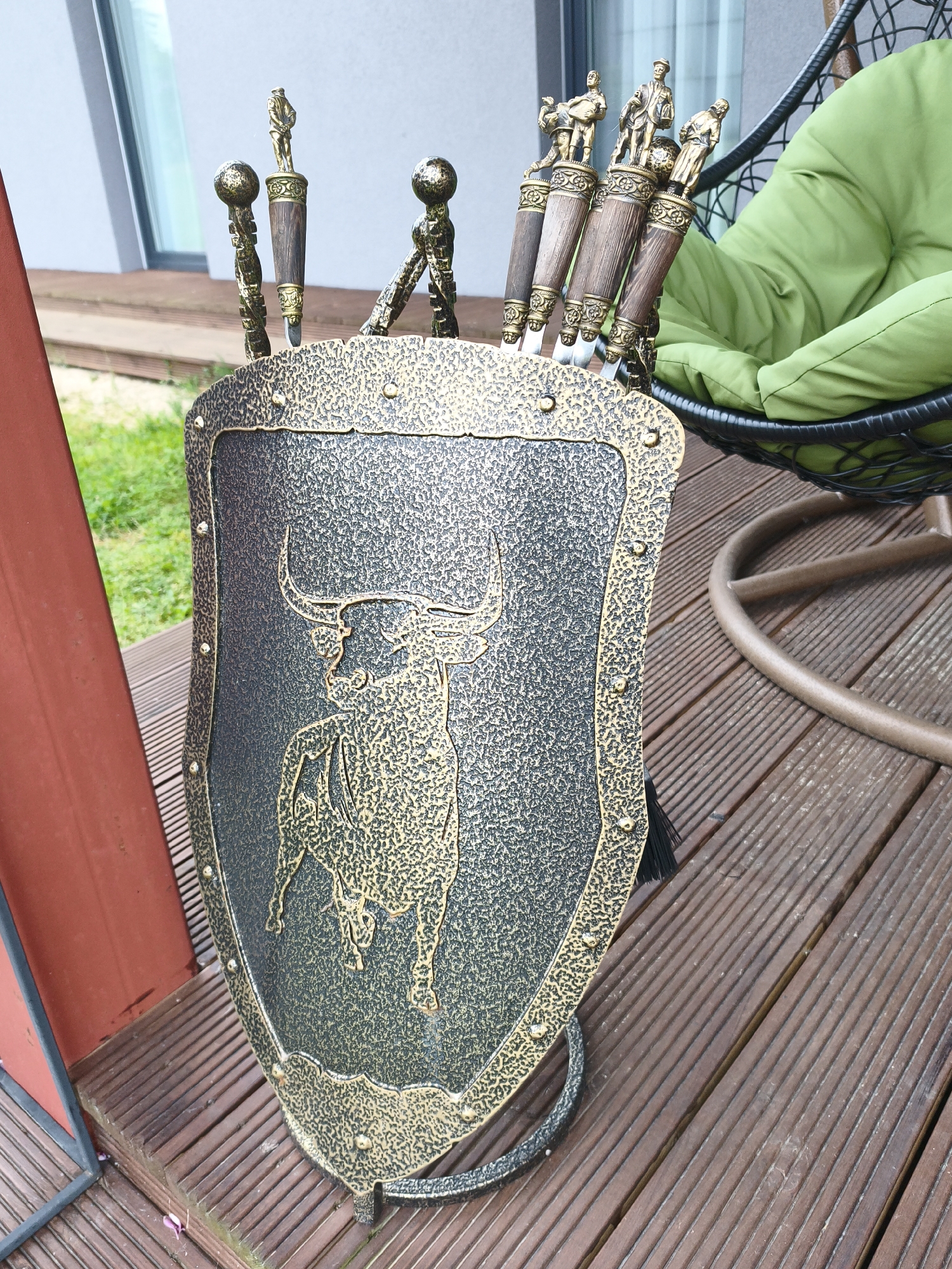 An ornamental shield, featuring a bull embossed in gold, stands on a wooden deck. The shield is dark gray with a textured, hammered finish and a gold border accented with gold studs. It is the centerpiece of a set of decorative items, with several skewers, each topped with a miniature statue of a man and a round finial, inserted into the top of the shield. A black tassel hangs from the bottom right side of the shield. The deck is made of dark brown wooden planks. In the background, a green cushion sits in a black wicker chair, and a portion of a building with a large window is visible. The scene appears to be outdoors.