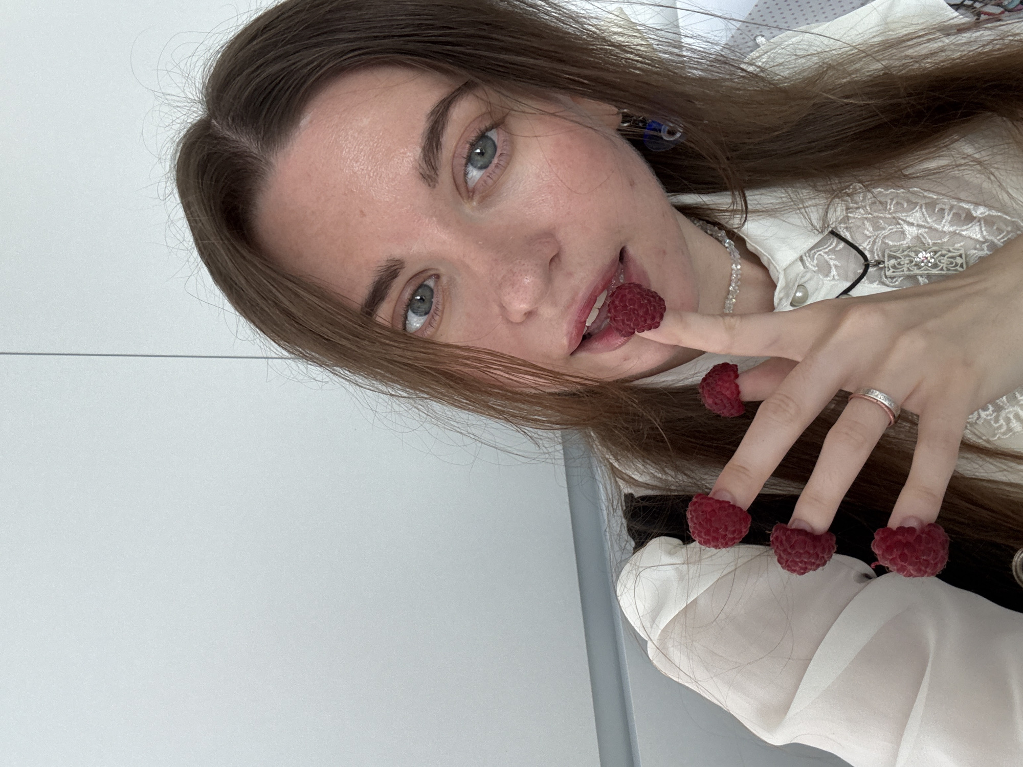 A young woman with brown hair and blue eyes is shown in a close-up, selfie-style shot. She is wearing a white blouse with lace detailing and a black vest. Several raspberries are placed on her fingertips and one is near her lips. She is also wearing a silver ring on her left hand and a necklace. The background appears to be a plain, light-colored wall with some vertical lines. The setting is indoors, likely an apartment or office.